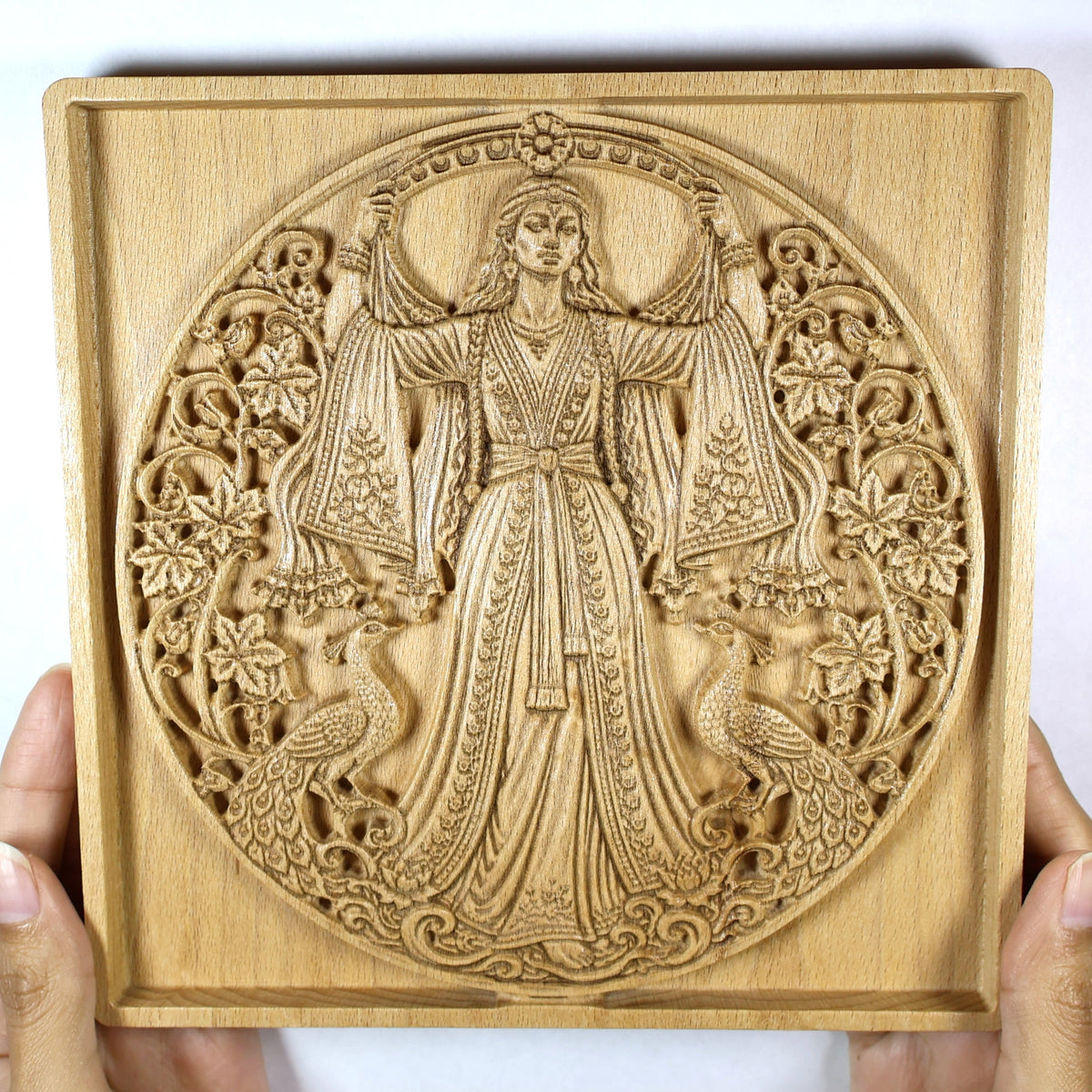 Anahita wood relief held in hands for size reference, showing the square frame, goddess carving, peacocks, and natural beech wood.
