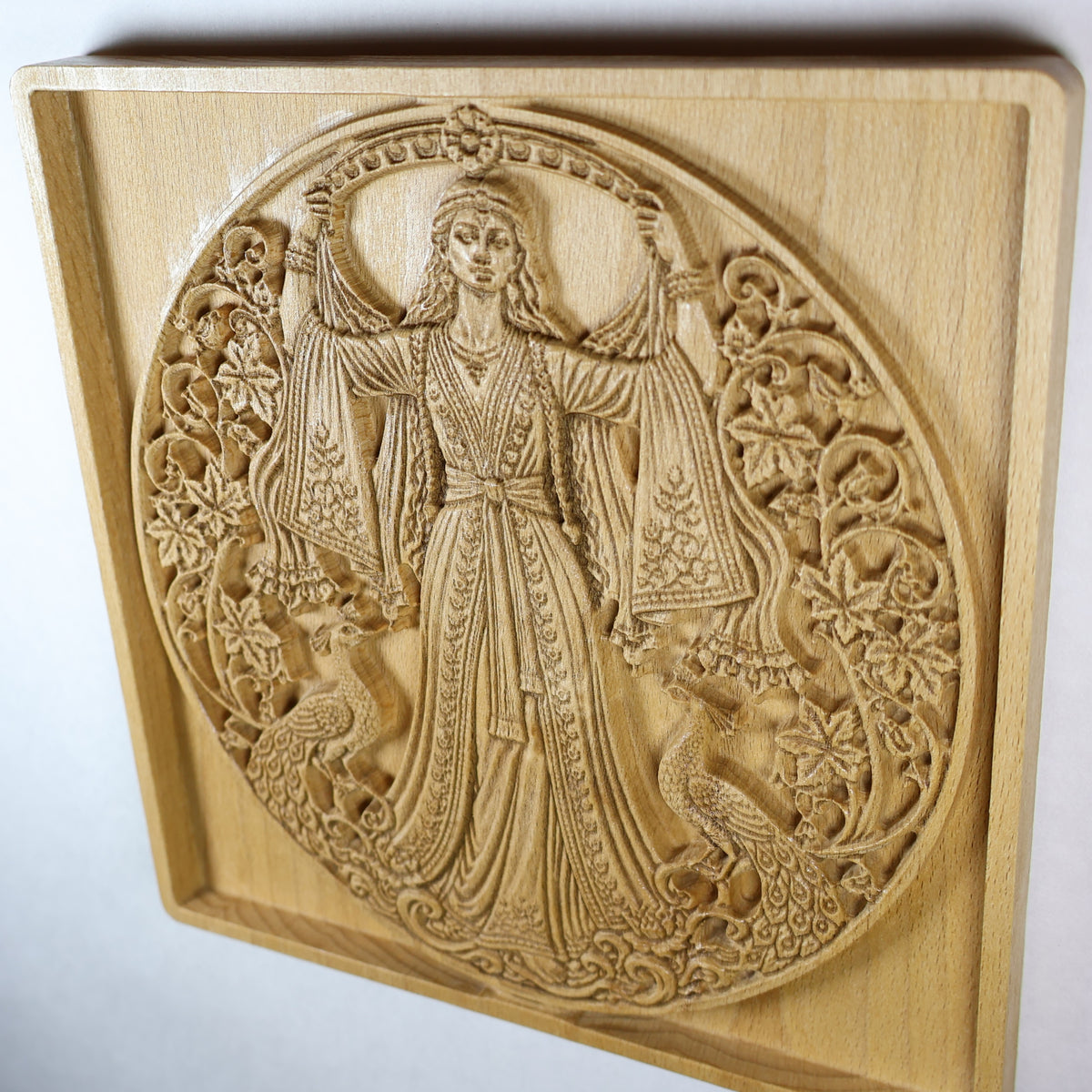 Angled view of the Anahita carving showing the goddess, ornate robe, peacocks, flowers, dimensional relief, and warm wood grain.