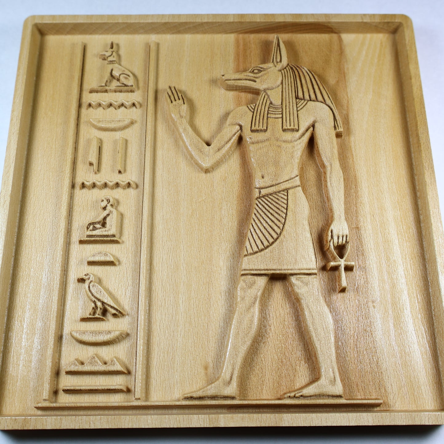 Angled view of an Anubis wood relief showing the carved jackal head, ankh, hieroglyphic column, raised details, and natural wood grain.