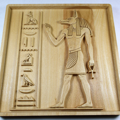 Angled view of an Anubis wood relief showing the carved jackal head, ankh, hieroglyphic column, raised details, and natural wood grain.