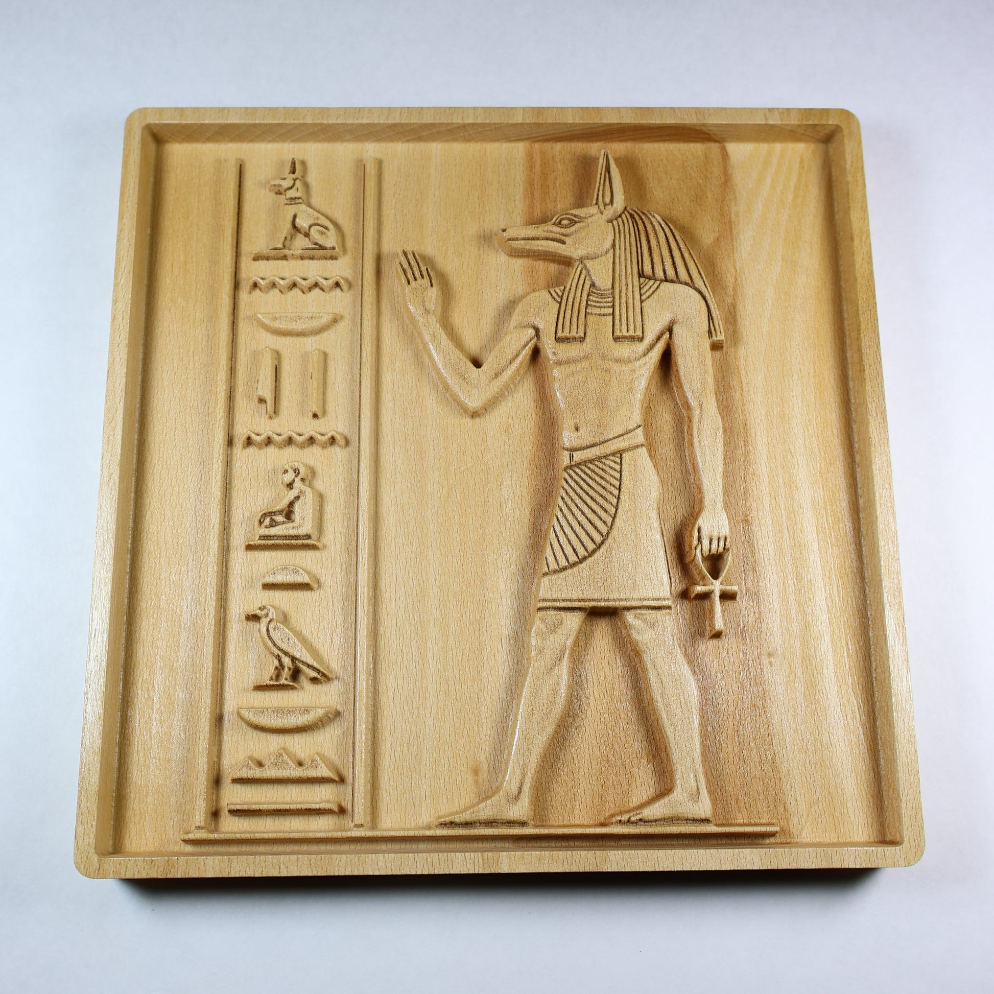 Low angle view of an Anubis wood relief showing the standing Egyptian figure, carved symbols, frame depth, and natural wood finish.