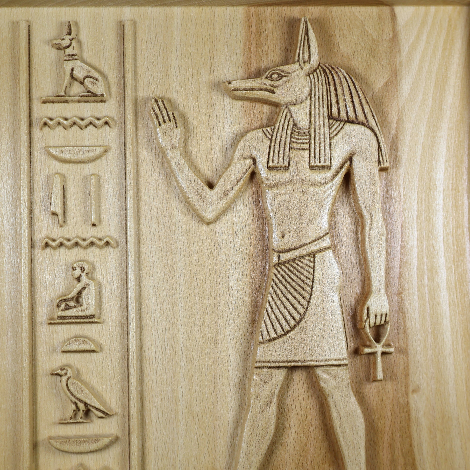 Macro view of an Anubis wood relief showing the carved jackal head, headdress, ankh, hieroglyphic symbols, and natural wood texture.