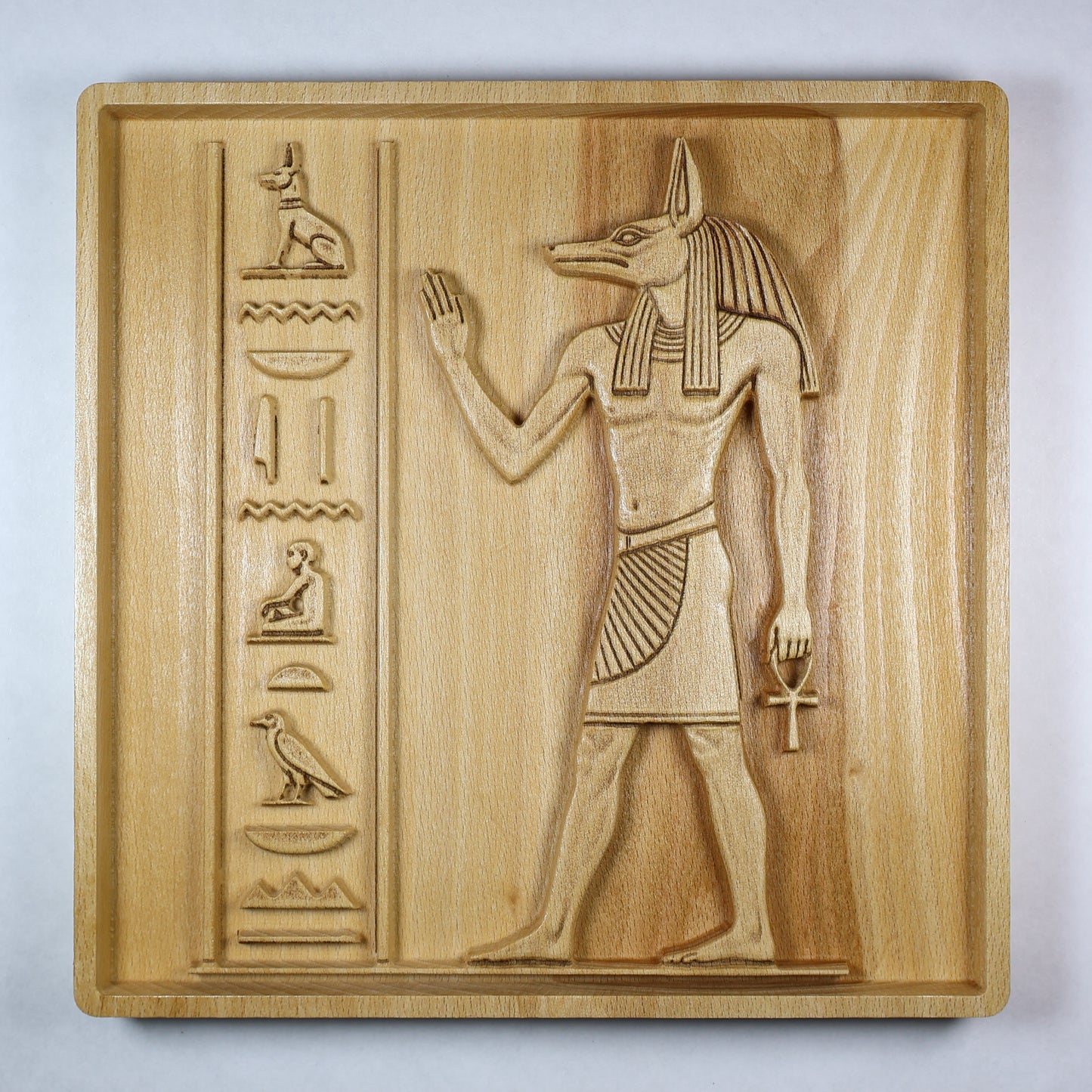 Straight-on detail of an Anubis wood relief showing the jackal-headed figure, ankh, raised hand, carved symbols, and natural wood grain.