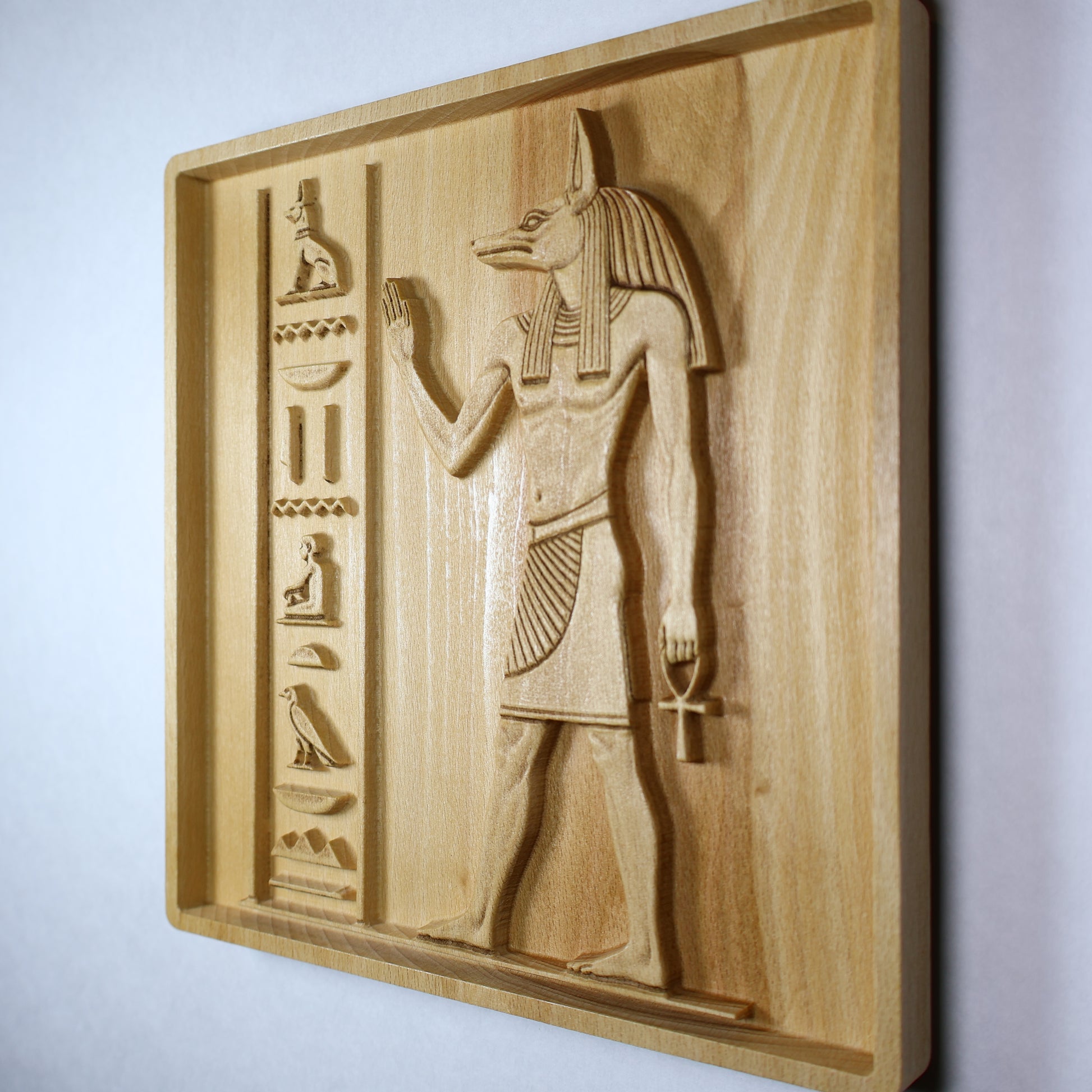 Right side angle of an Anubis wood relief showing the raised Egyptian figure, hieroglyphic carvings, square frame, and natural wood finish.