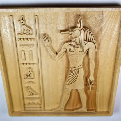 Close angled detail of an Anubis wood relief showing the jackal head, raised arm, carved symbols, relief texture, and natural wood grain.