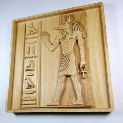 Three-quarter view of an Anubis wood relief showing the Egyptian figure, ankh, hieroglyphic column, recessed frame, and natural wood finish.