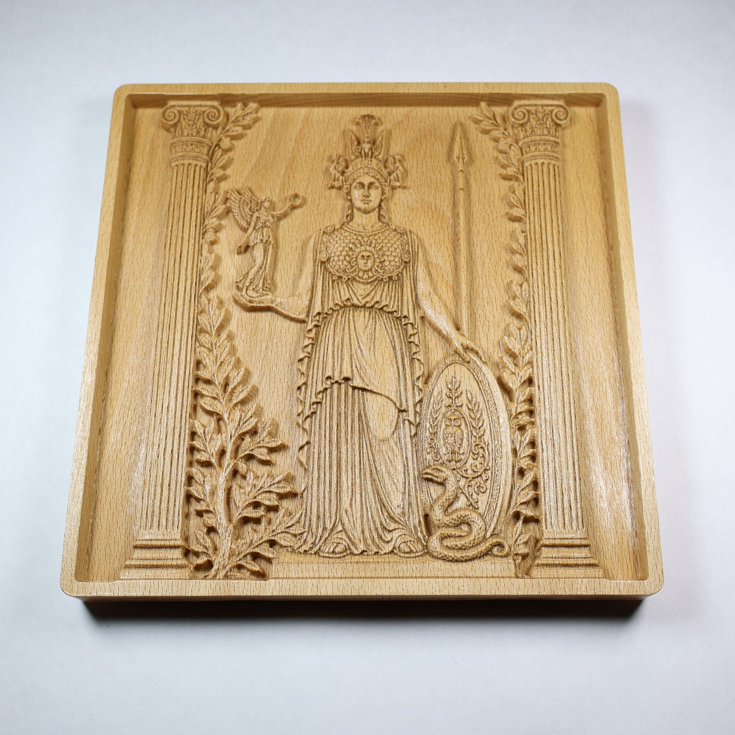 Low angled view of an Athena wood carving showing the goddess, shield, spear, layered robe details, raised relief, and natural wood grain.