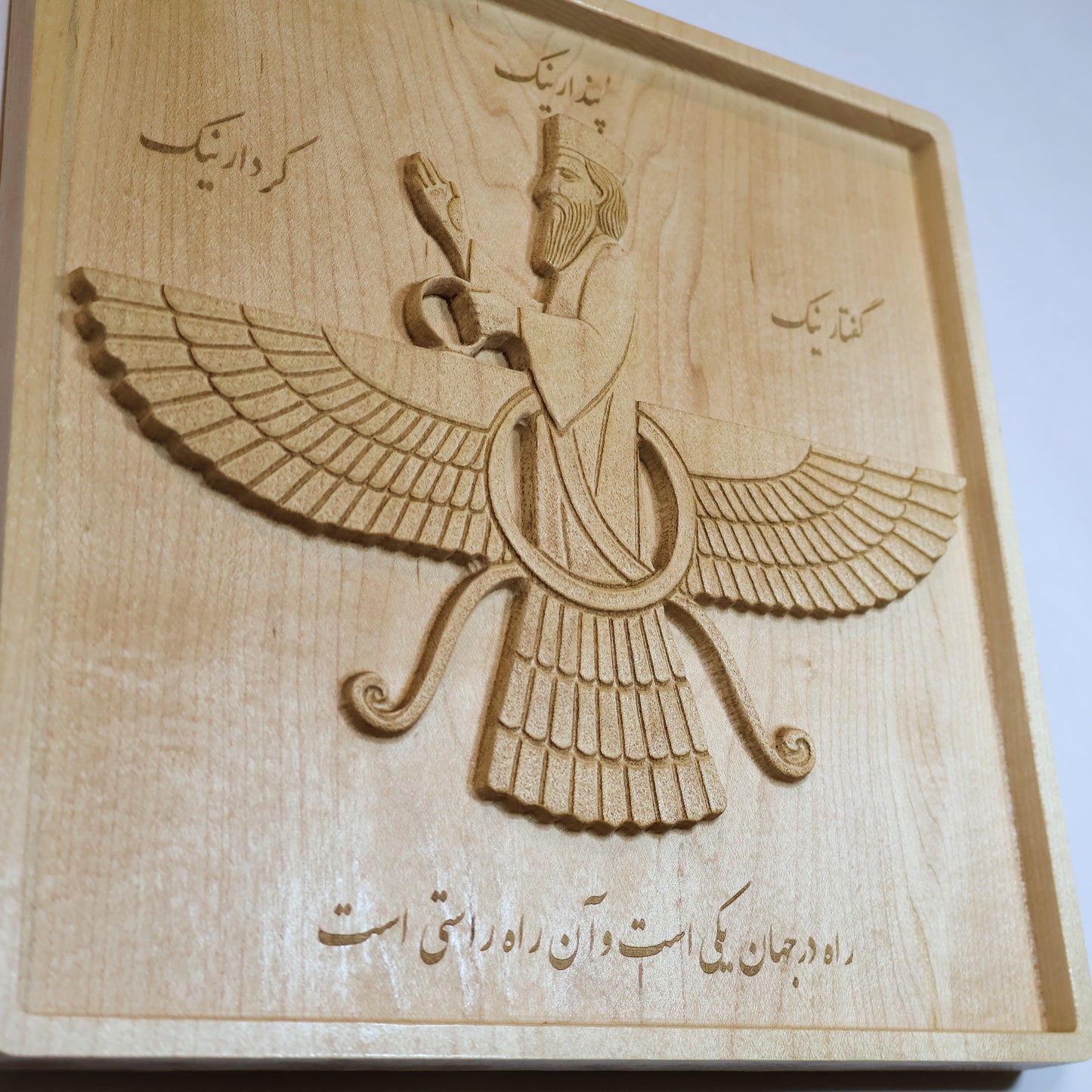 Low perspective of the Faravahar wood relief showing the figure, feather carving, inscriptions, raised surfaces, and solid maple panel.
