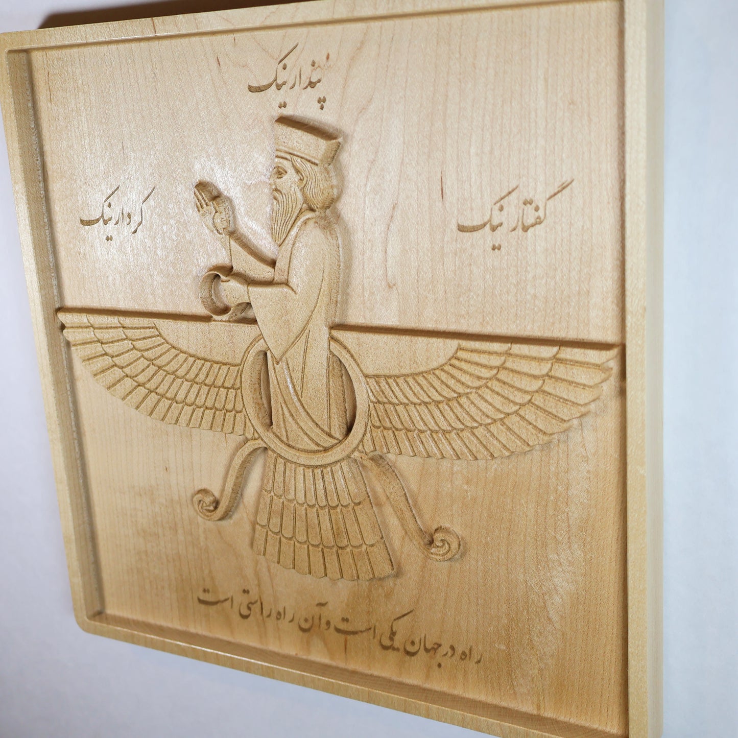 Angled view of the Faravahar relief showing the winged figure, Farsi writing, layered feathers, recessed frame, and carved hard maple depth.
