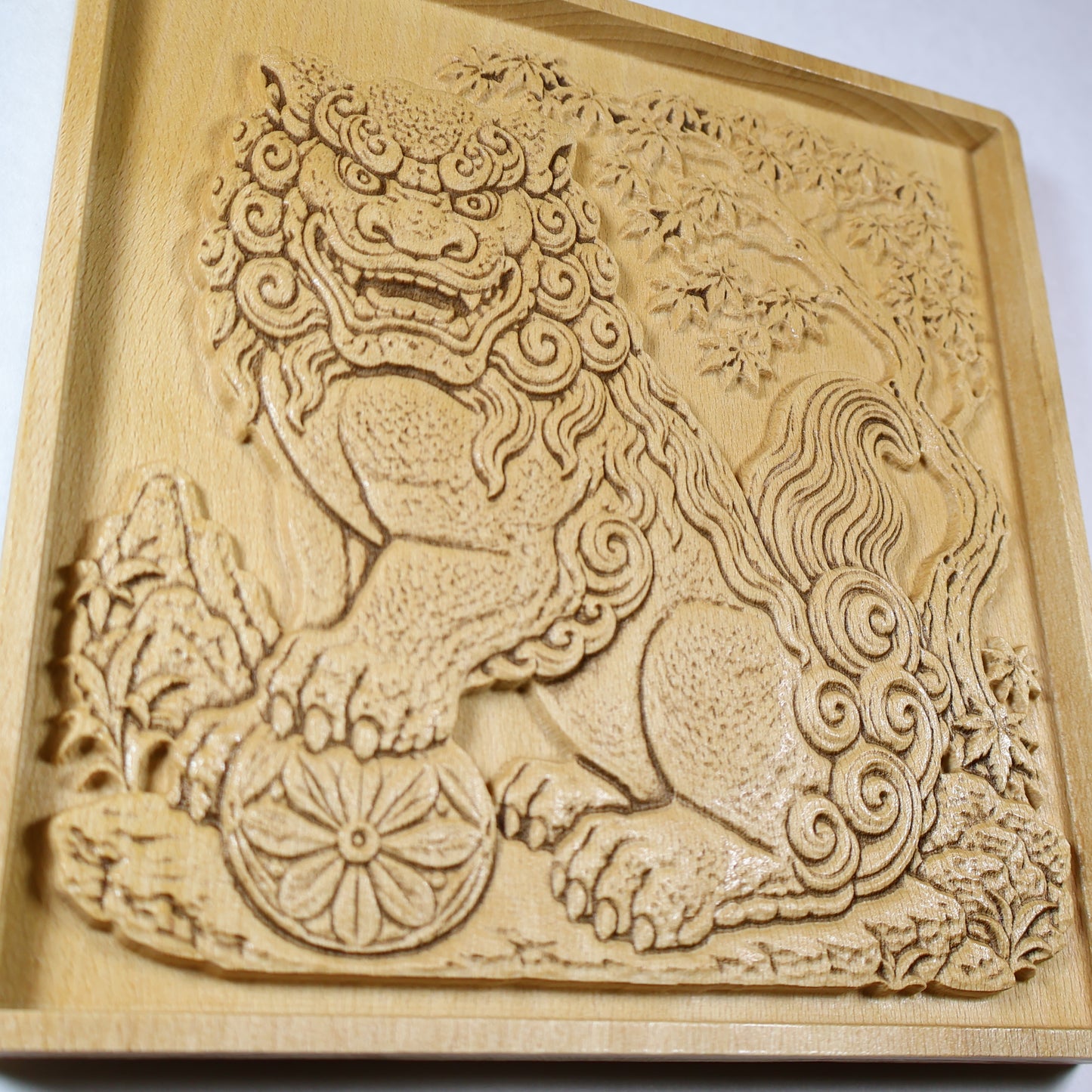 Close angled view of a Japanese Komainu guardian lion dog wood relief with a carved floral ball, curled mane, flowing tail, maple tree, and natural wood finish.