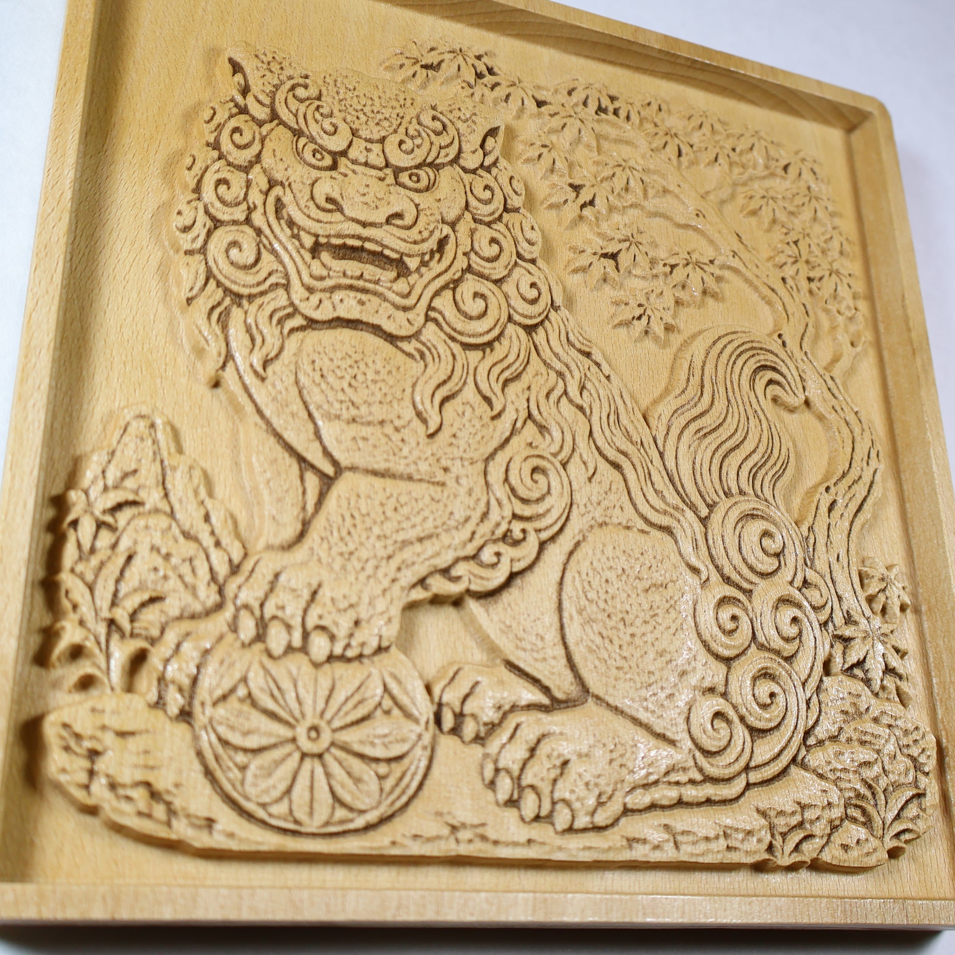 Close angled view of a Japanese Komainu guardian lion dog wood relief with a carved floral ball, curled mane, flowing tail, maple tree, and natural wood finish.