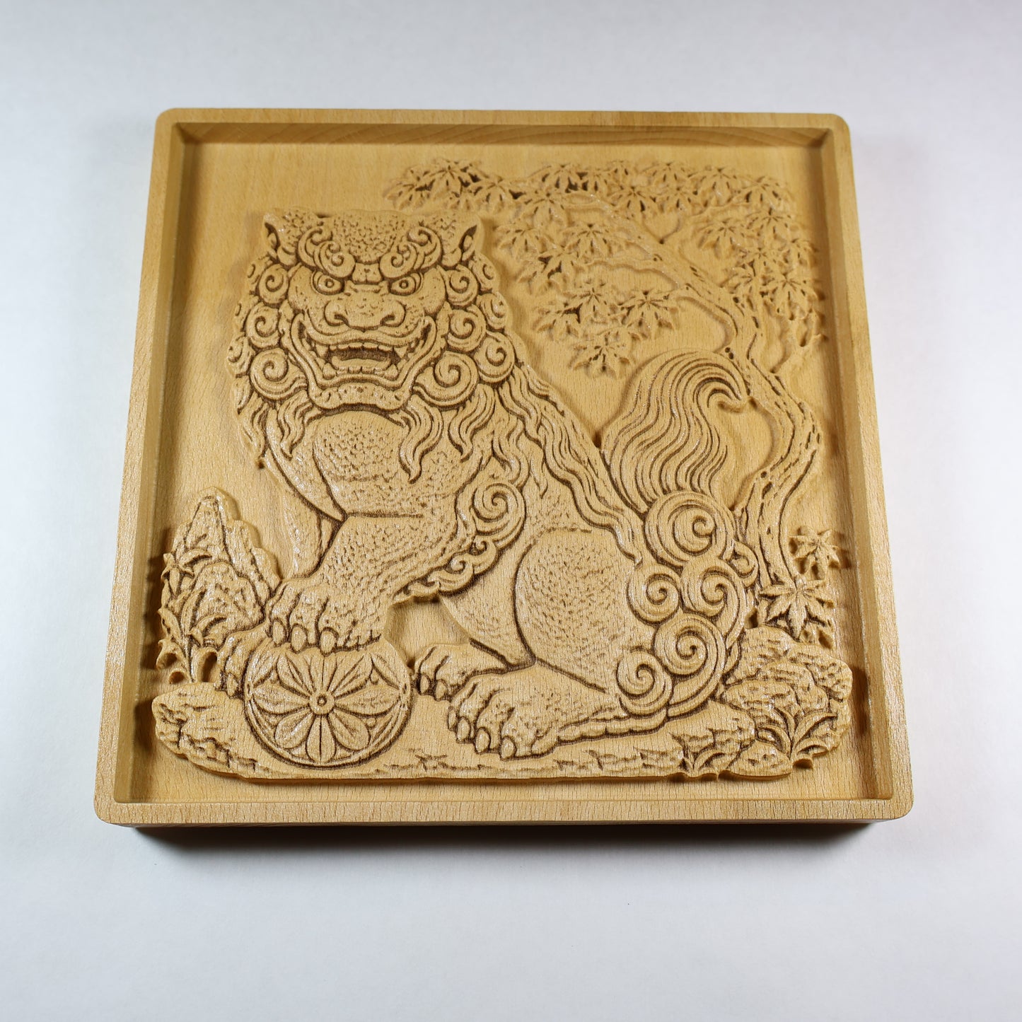 Angled view of a Japanese Komainu guardian lion dog wood relief with a carved floral ball, curled mane, maple tree background, square frame, and natural wood finish.