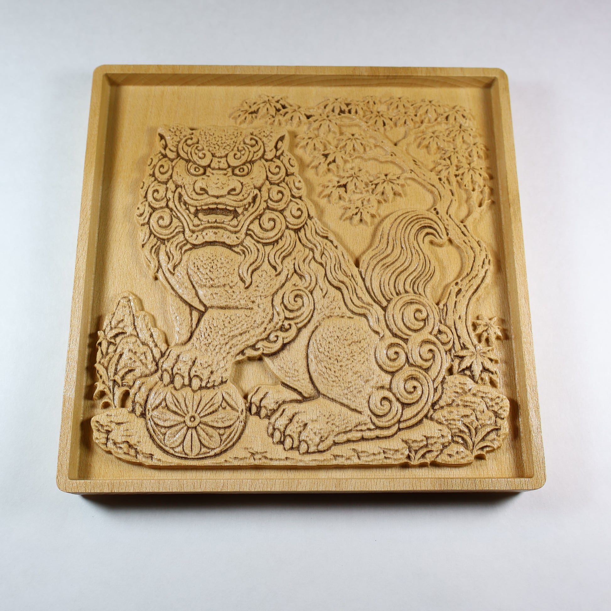 Angled view of a Japanese Komainu guardian lion dog wood relief with a carved floral ball, curled mane, maple tree background, square frame, and natural wood finish.