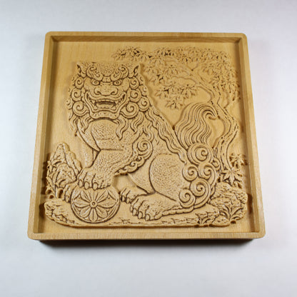 Angled view of a Japanese Komainu guardian lion dog wood relief with a carved floral ball, curled mane, maple tree background, square frame, and natural wood finish.