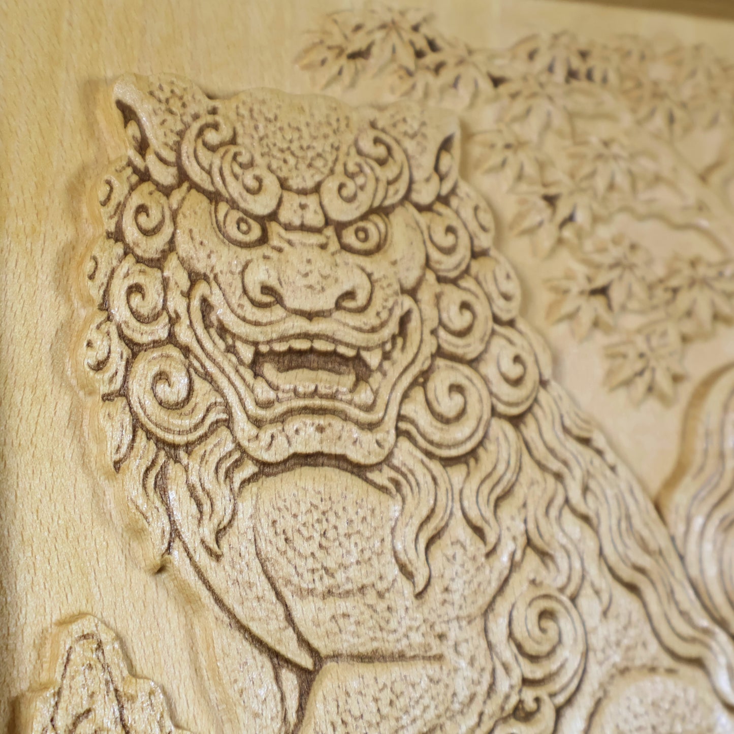 Close-up of a Japanese Komainu guardian lion dog wood relief showing the carved face, open mouth, curled mane, maple tree background, and natural wood finish.