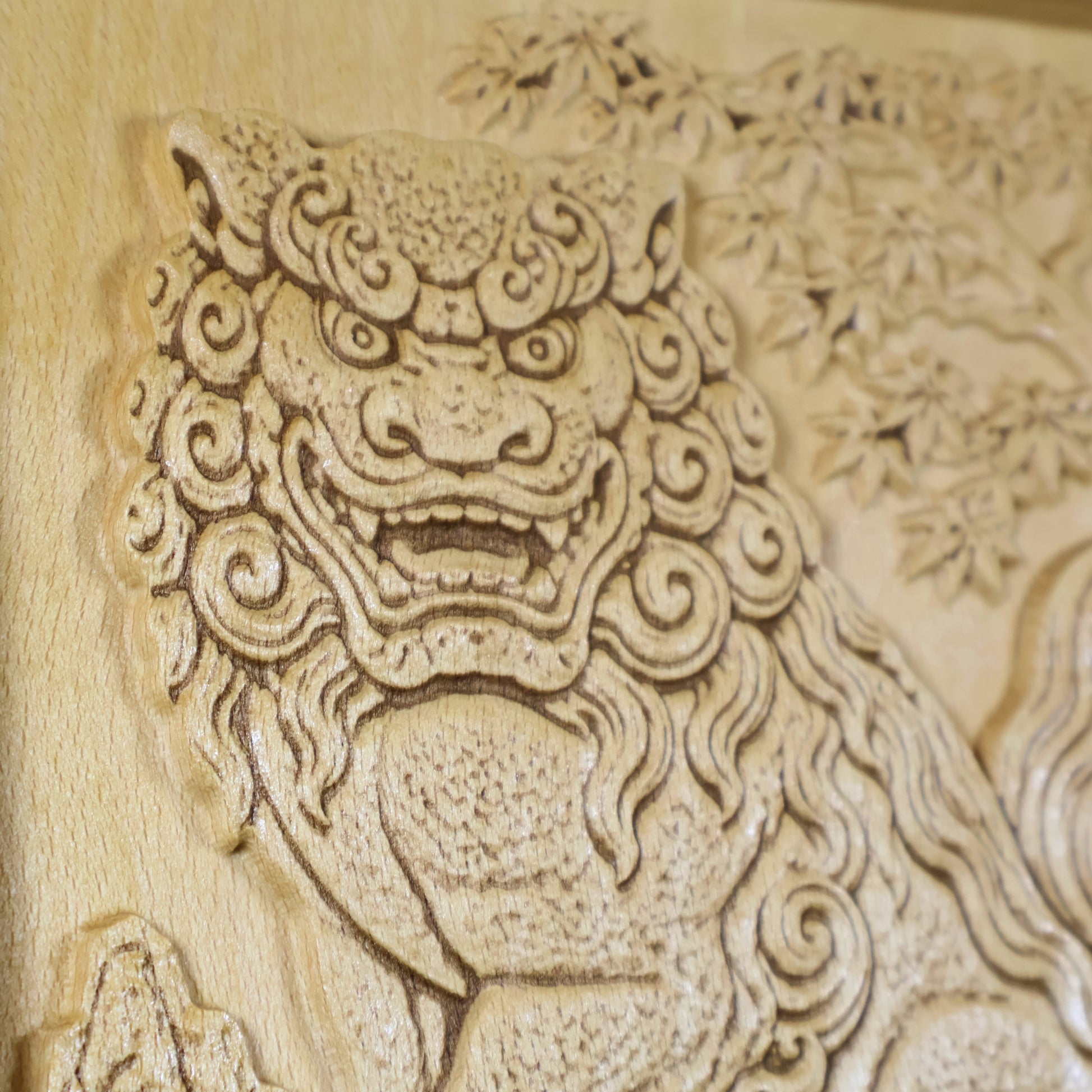 Close-up of a Japanese Komainu guardian lion dog wood relief showing the carved face, open mouth, curled mane, maple tree background, and natural wood finish.