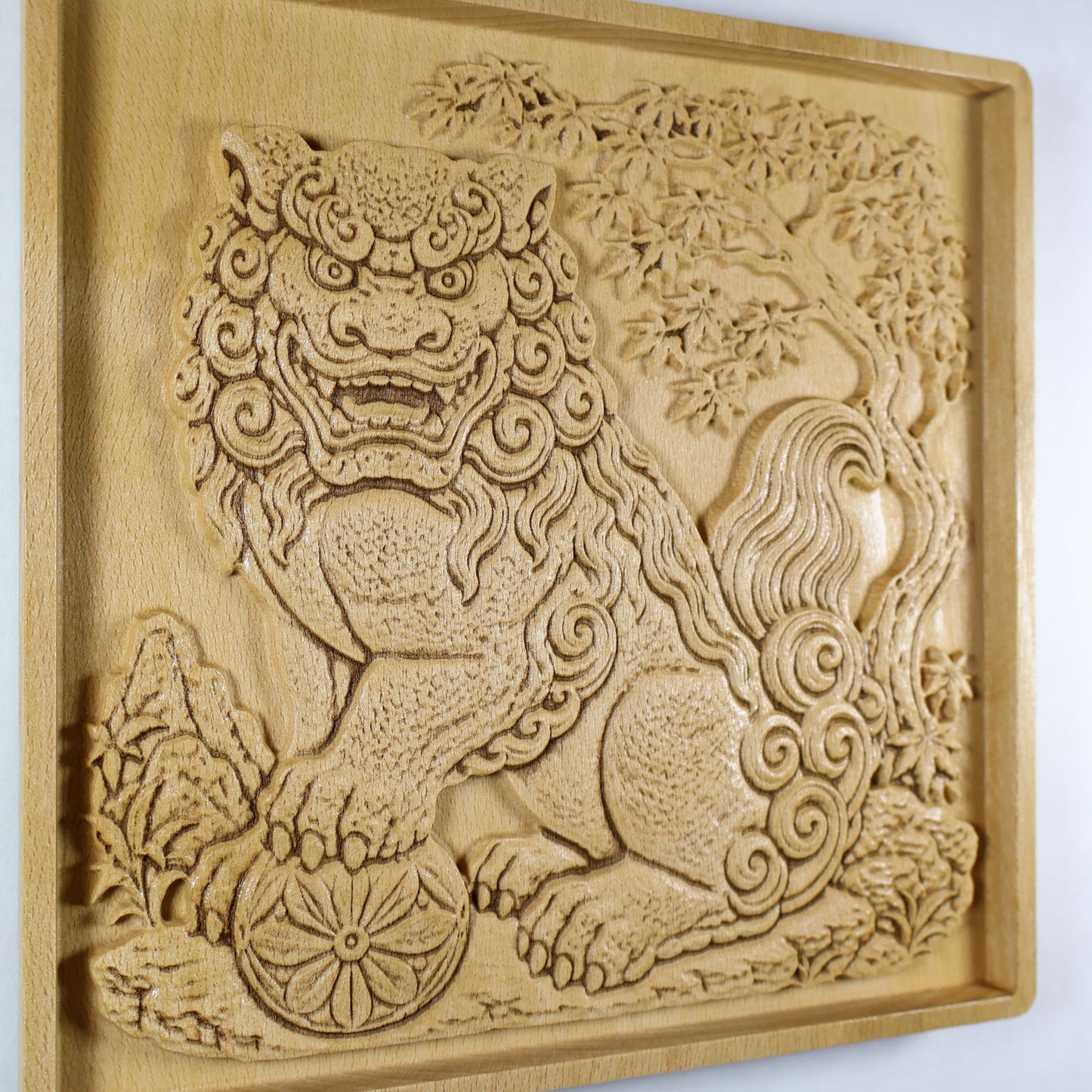 Angled close-up of a Japanese Komainu guardian lion dog wood relief showing the carved guardian, floral ball, curled mane and tail, maple tree, and natural wood finish.