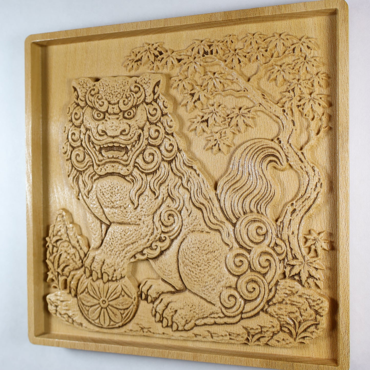 Angled side view of a Japanese Komainu guardian lion dog wood relief showing the carved guardian, floral ball, curled mane and tail, maple tree branches, and natural wood finish.