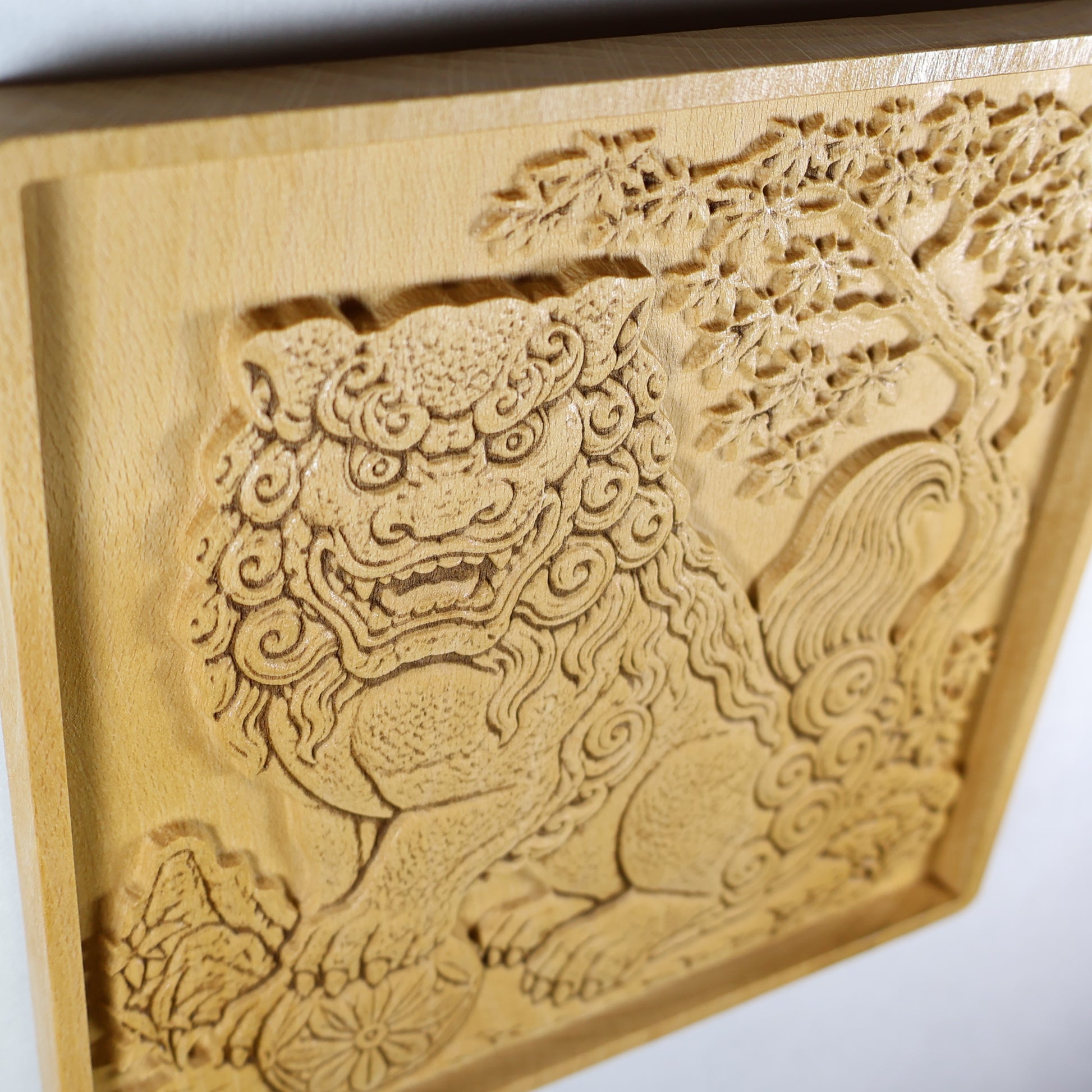 Low side-angle view of a Japanese Komainu wood wall art panel showing raised carving depth, shadowed details, maple branches, curled tail, and polished natural wood.
