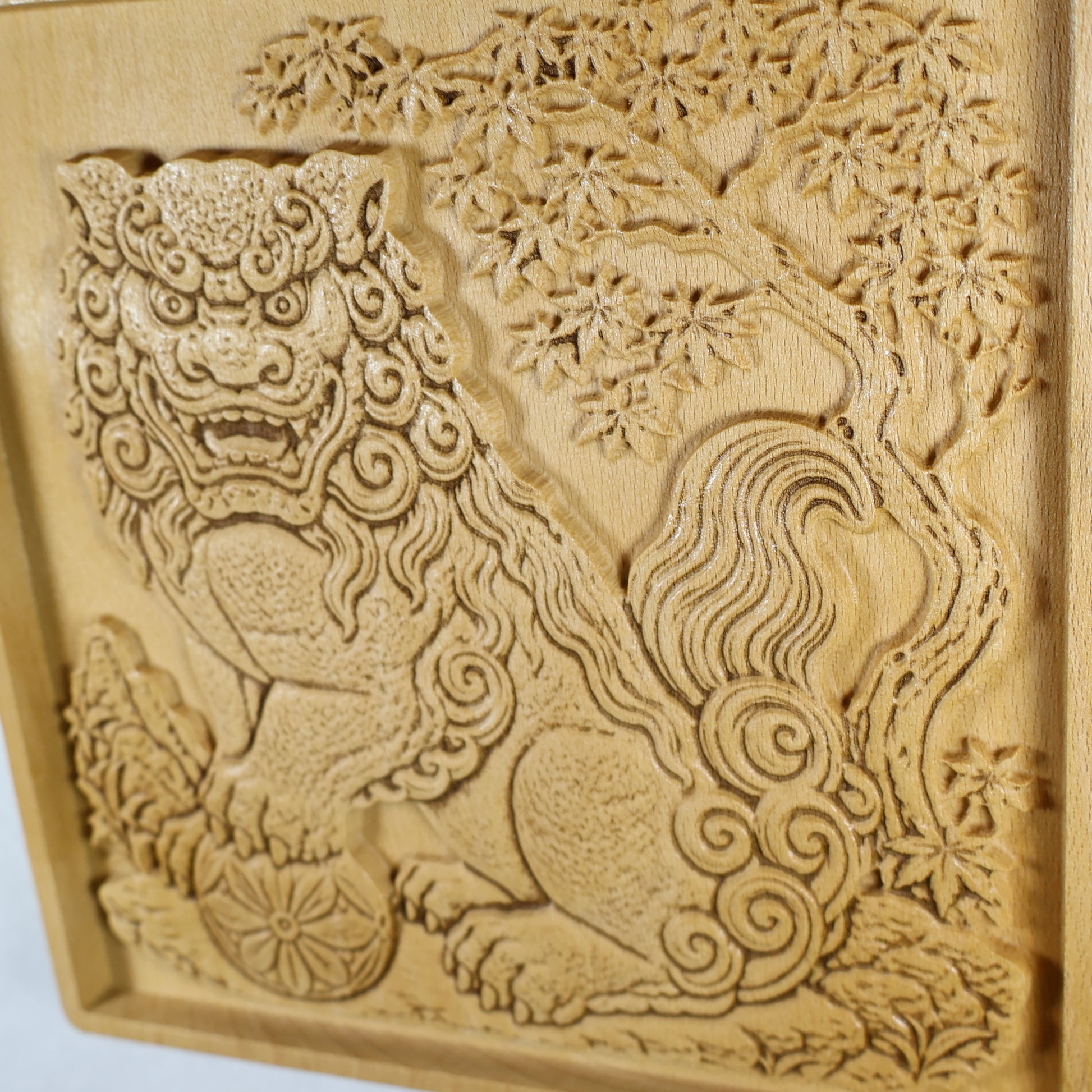 Close side detail of a Japanese Komainu wood relief showing the guardian lion, carved maple tree, flowing tail scrolls, flower ball, and raised natural wood texture.