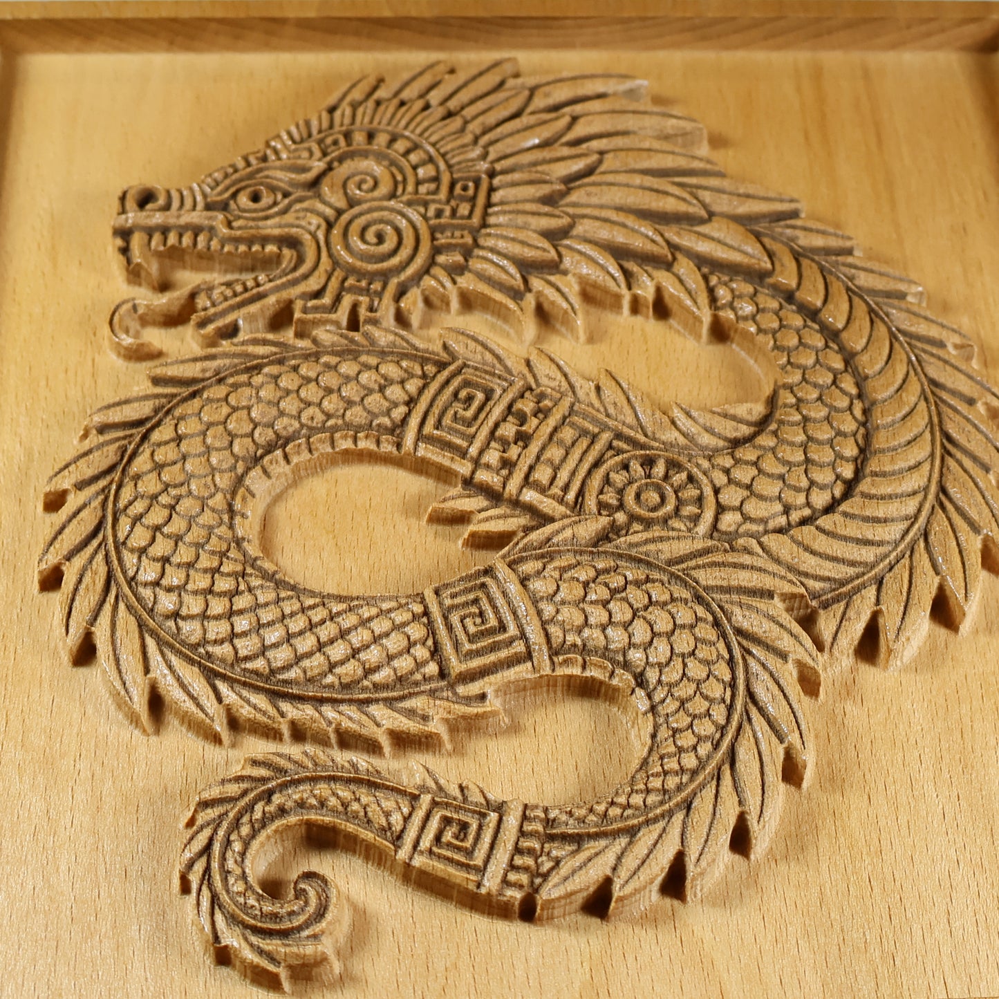 Macro detail of Quetzalcoatl wood carving showing serpent head with open jaw, spiral eye motif, layered feather plumes, interlocking scales, and laser-engraved line work on solid beech wood