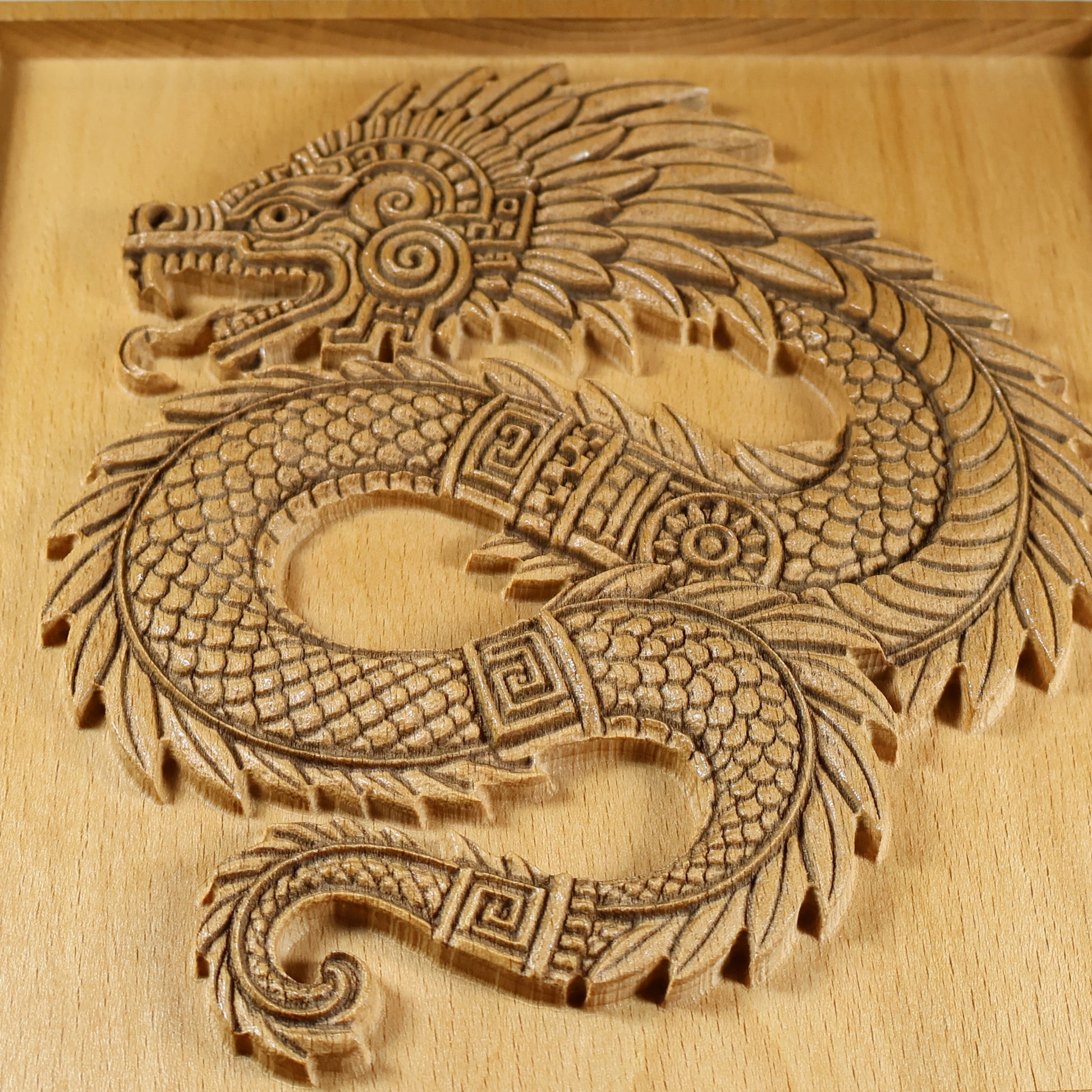 Macro detail of Quetzalcoatl wood carving showing serpent head with open jaw, spiral eye motif, layered feather plumes, interlocking scales, and laser-engraved line work on solid beech wood