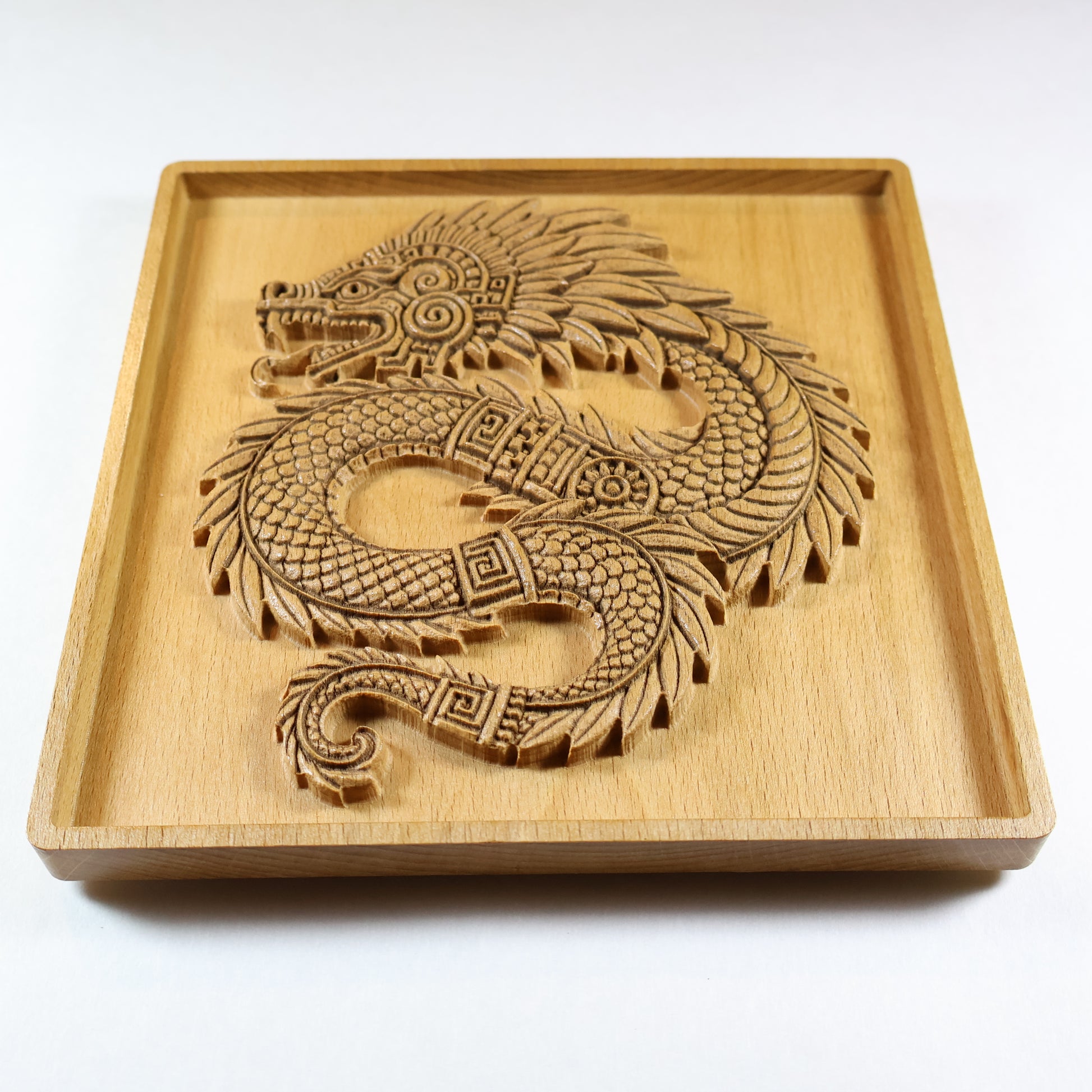 Low-angle close-up of Quetzalcoatl beech wood relief showing dramatic 3D carved depth, coiled feathered serpent body, layered scale texture, and spiral tail detail, 7 inch Aztec wood wall plaque