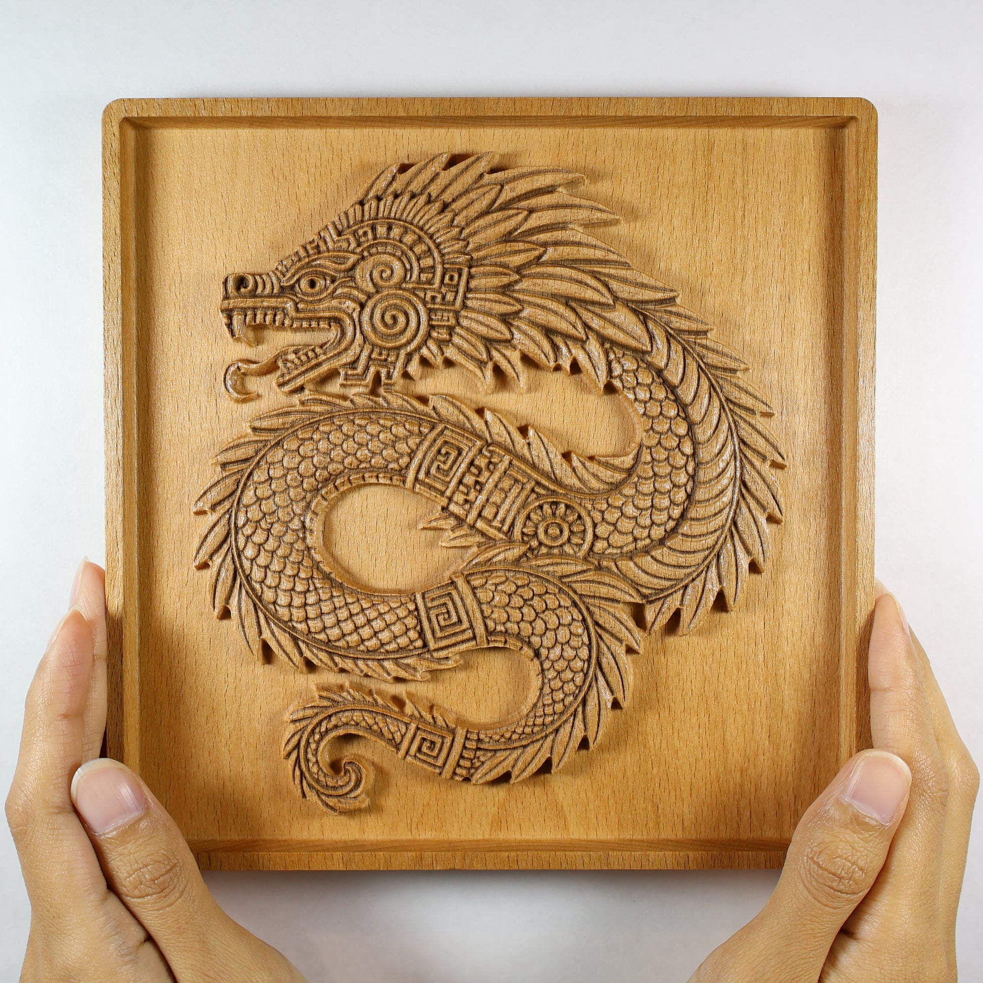 Person holding Quetzalcoatl 7x7 inch beech wood wall art with both hands, showing full coiled feathered serpent relief, open jaw, plume crest, reptile-scale body, geometric Aztec bands, curled tail, and recessed border frame, size reference
