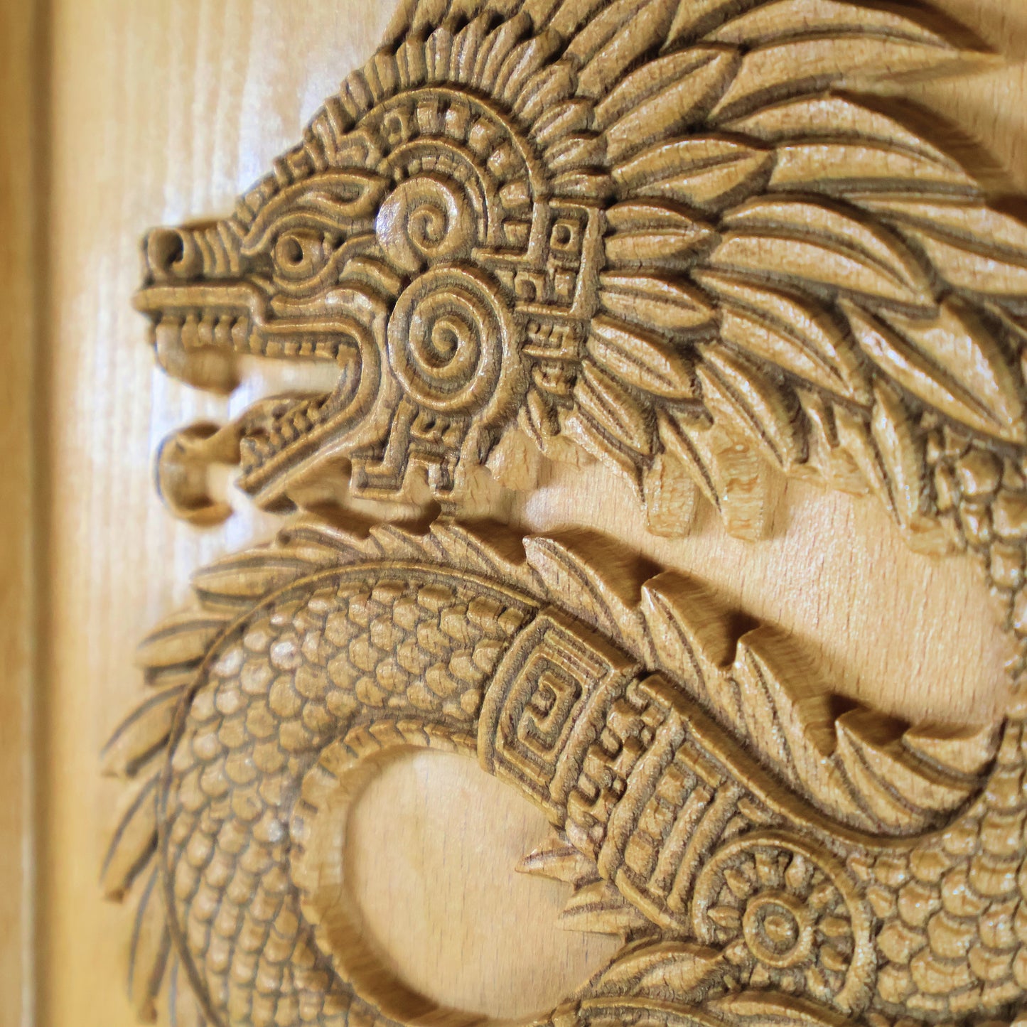 Angled close-up of Quetzalcoatl beech wood relief showing dramatic carved relief depth, feathered serpent head with spiral ornament, layered feather plumes, fish-scale body texture, and geometric Aztec patterning