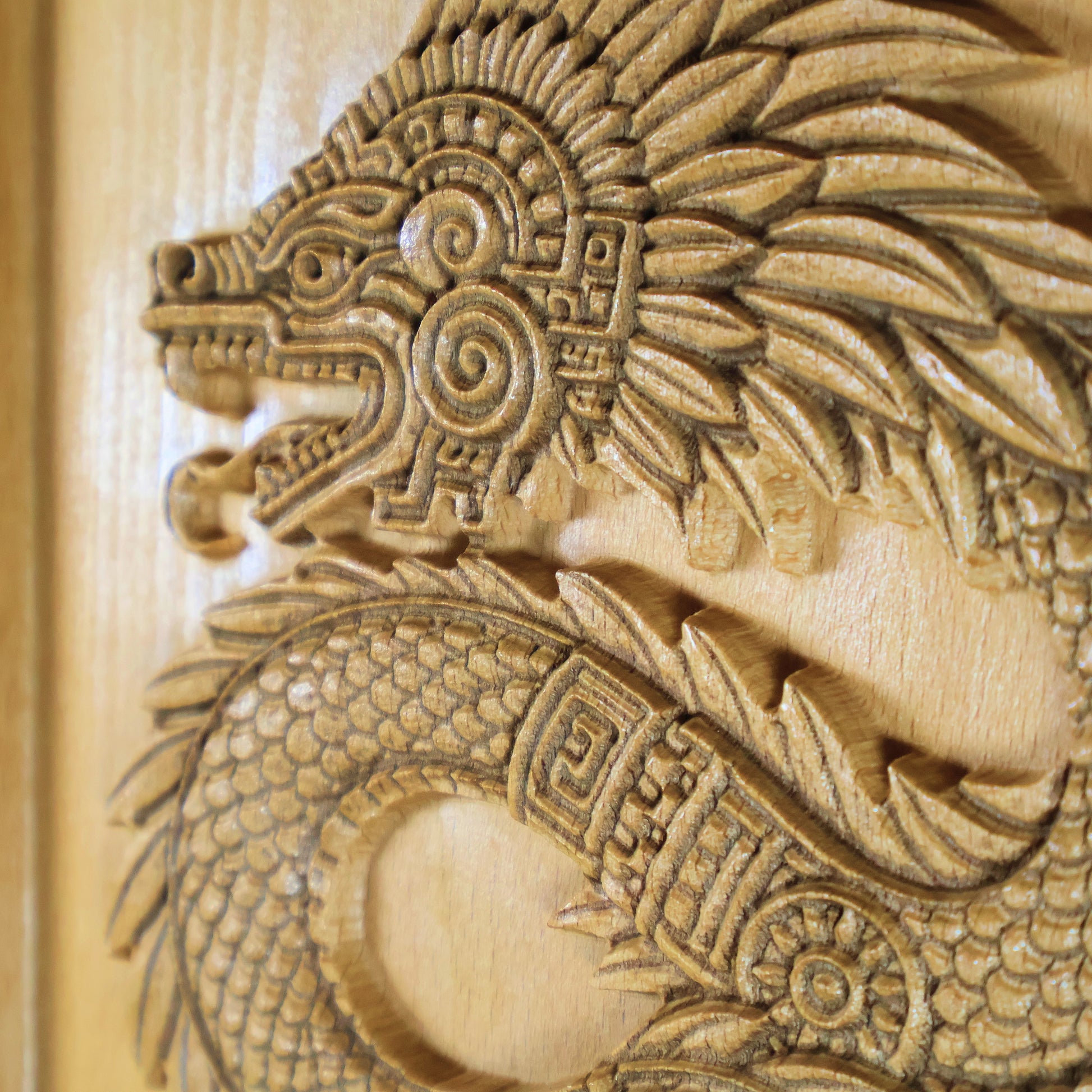 Angled close-up of Quetzalcoatl beech wood relief showing dramatic carved relief depth, feathered serpent head with spiral ornament, layered feather plumes, fish-scale body texture, and geometric Aztec patterning