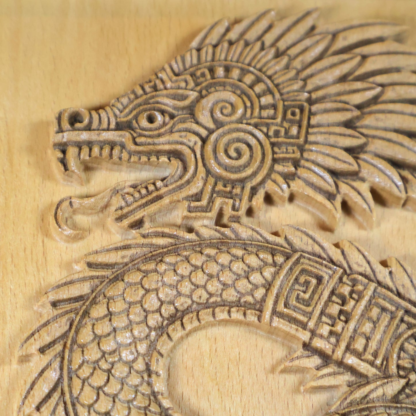 Extreme close-up of Quetzalcoatl serpent head carved in beech wood, revealing open fanged jaw, circular spiral cheek ornament, raised feather crest, geometric neck band, and fine CNC-engraved texture