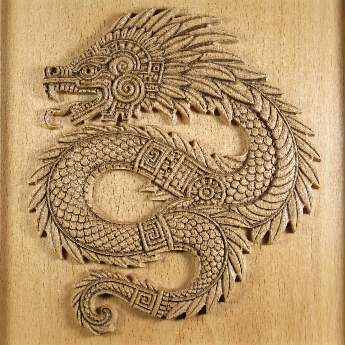 Full front detail of Quetzalcoatl carved beech wood relief, complete coiled feathered serpent with open jaw, spiral cheek disc, layered plume crest, fish-scale body, meander band collar, and curled tail on warm wood grain