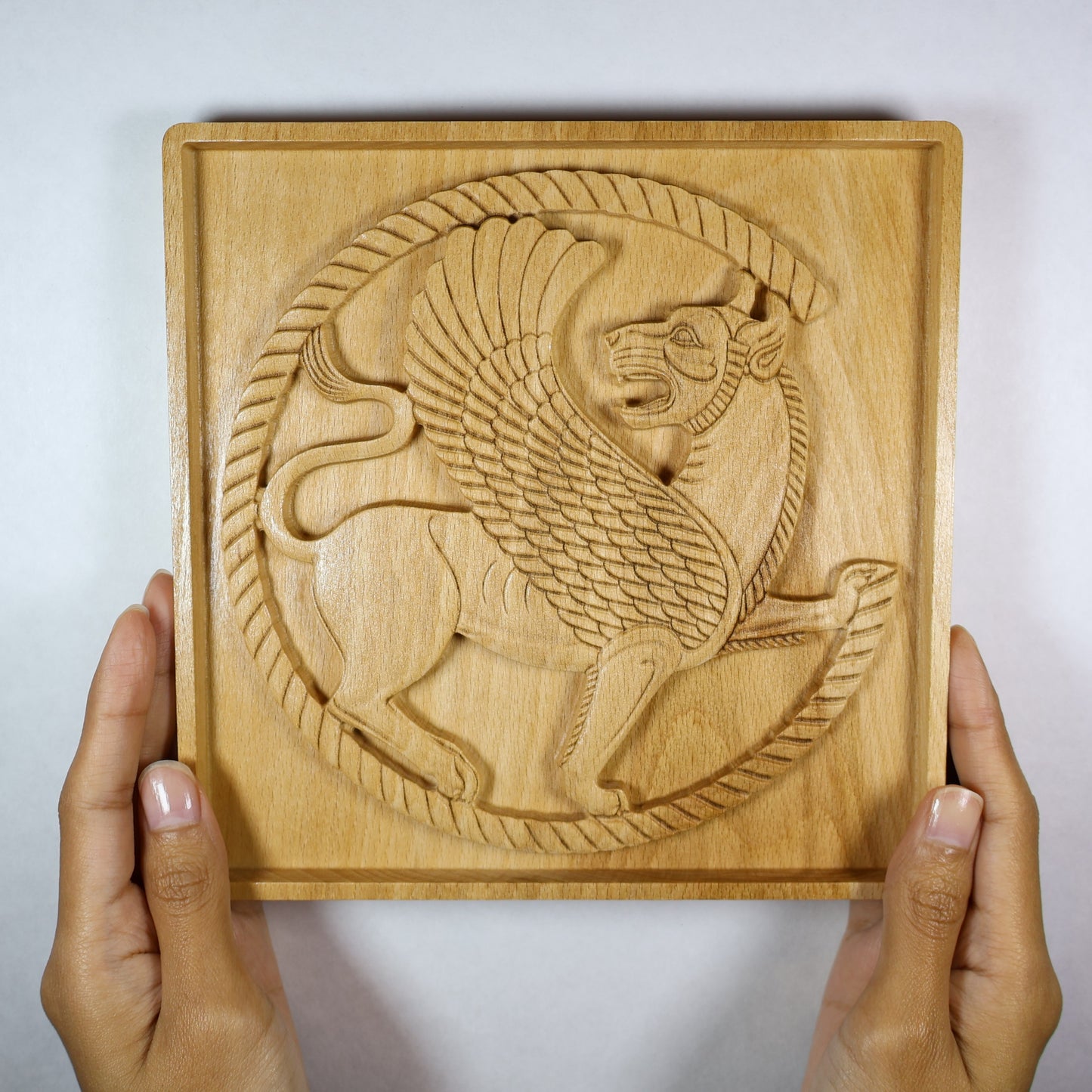 Achaemenid Winged Lion Roundel held in hands, showing the size, carved circular griffin design, square frame, and natural wood finish.
