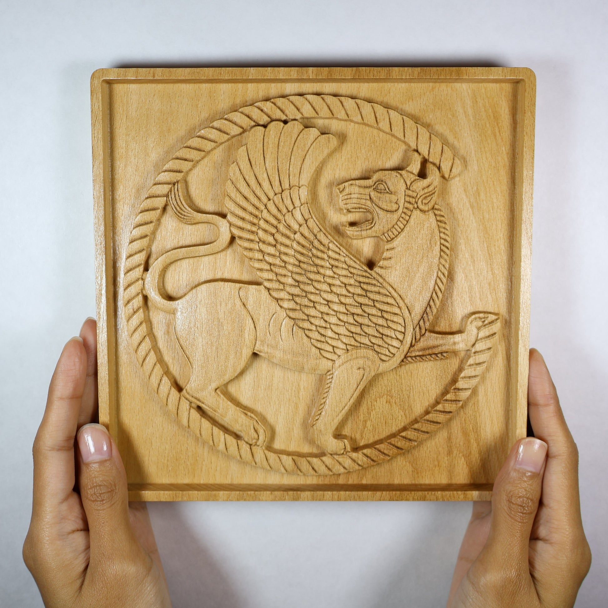 Achaemenid Winged Lion Roundel held in hands, showing the size, carved circular griffin design, square frame, and natural wood finish.