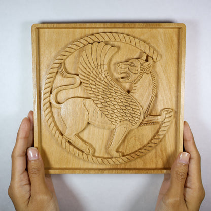 Achaemenid Winged Lion Roundel held in hands, showing the size, carved circular griffin design, square frame, and natural wood finish.