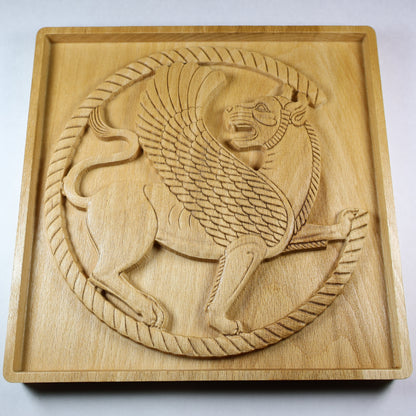 Low angled view of a Persian griffin wood relief showing the winged lion roundel, raised carving depth, feather details, and natural wood grain.