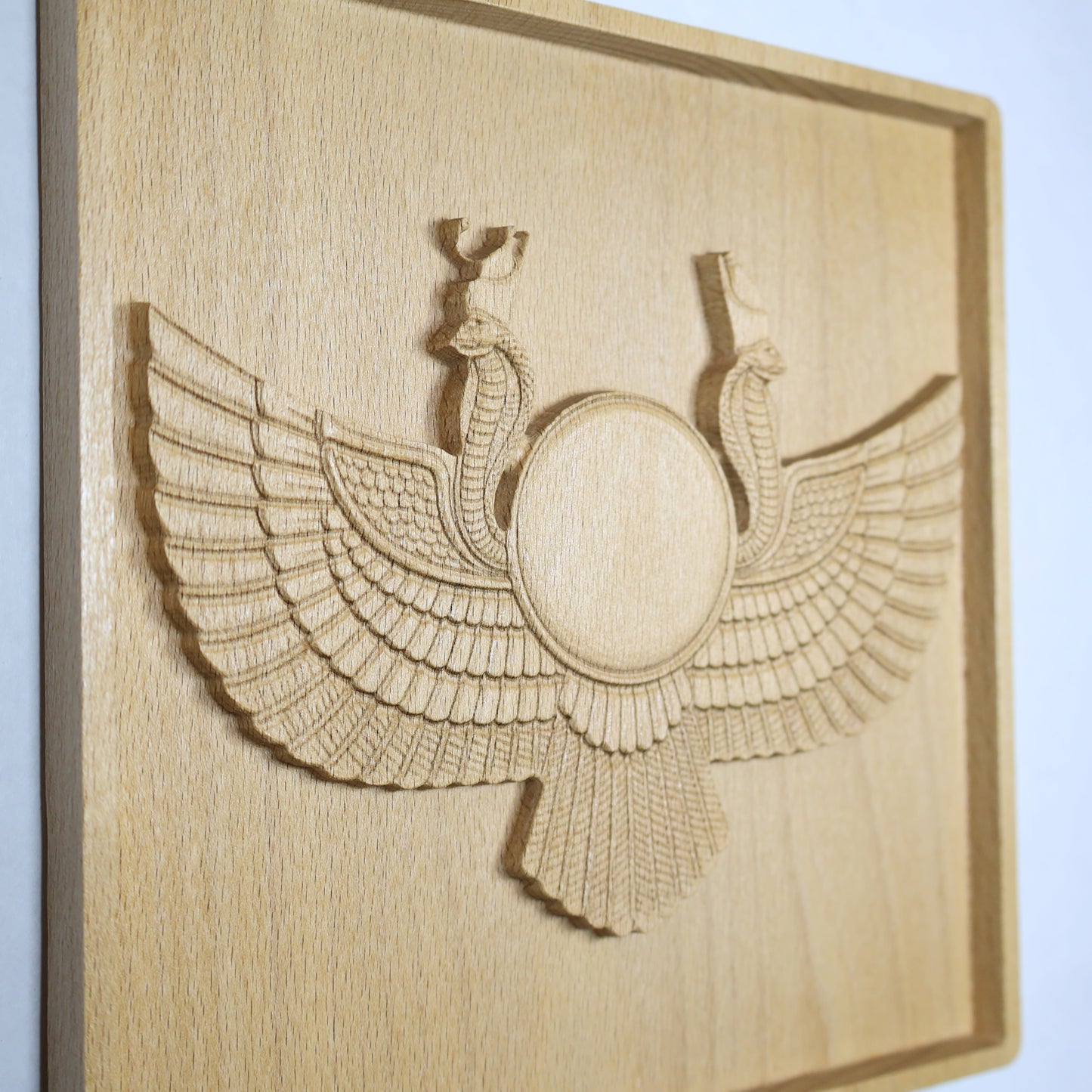 Side-angle view of an Egyptian Winged Sun Disk wood relief showing the raised sun disk, carved wings, twin cobras, feather details, and natural beech wood finish.