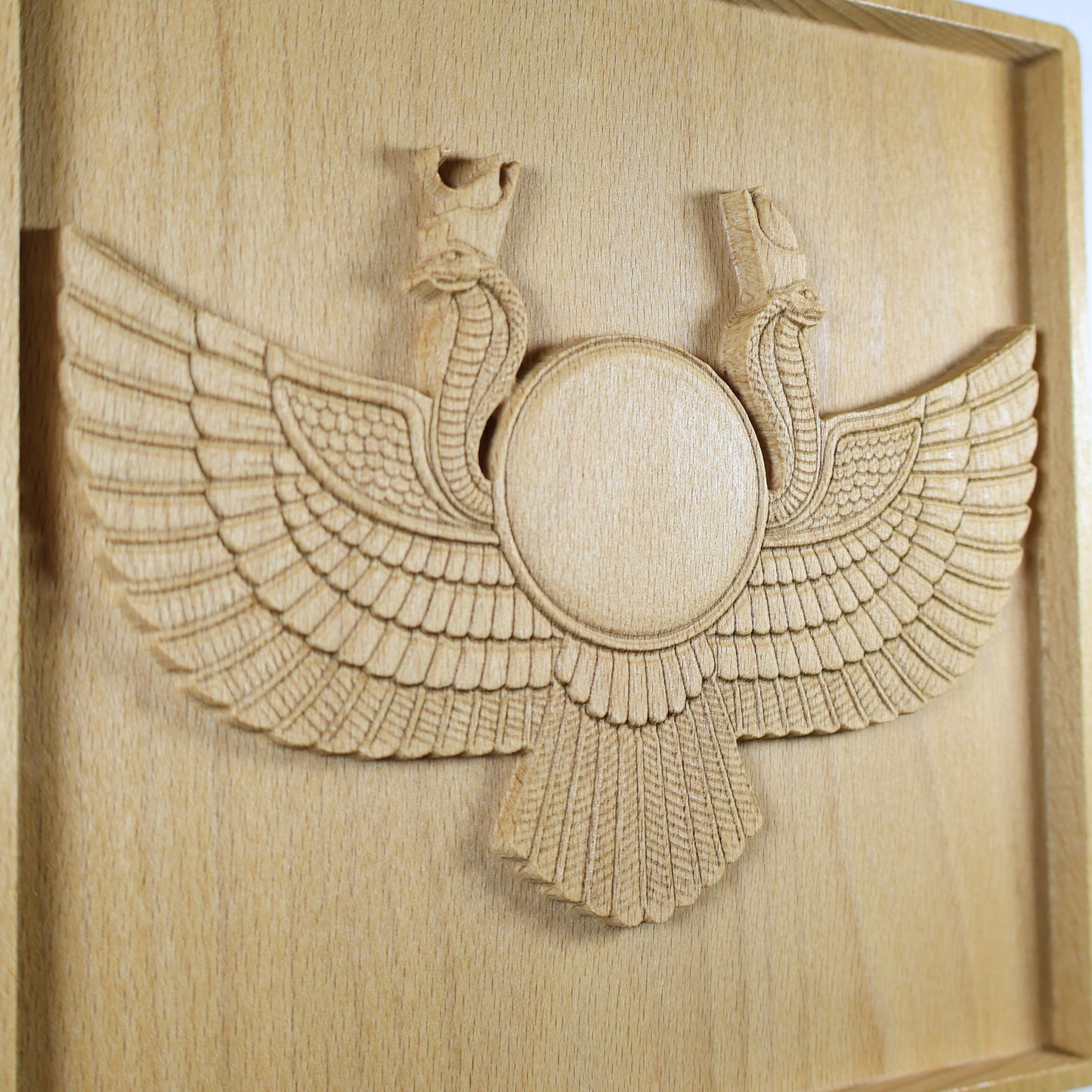 Angled close-up of an Egyptian Winged Sun Disk wood relief showing the Ra-Horus sun emblem, cobra forms, layered feathers, fan-shaped lower wing, and natural beech wood finish.