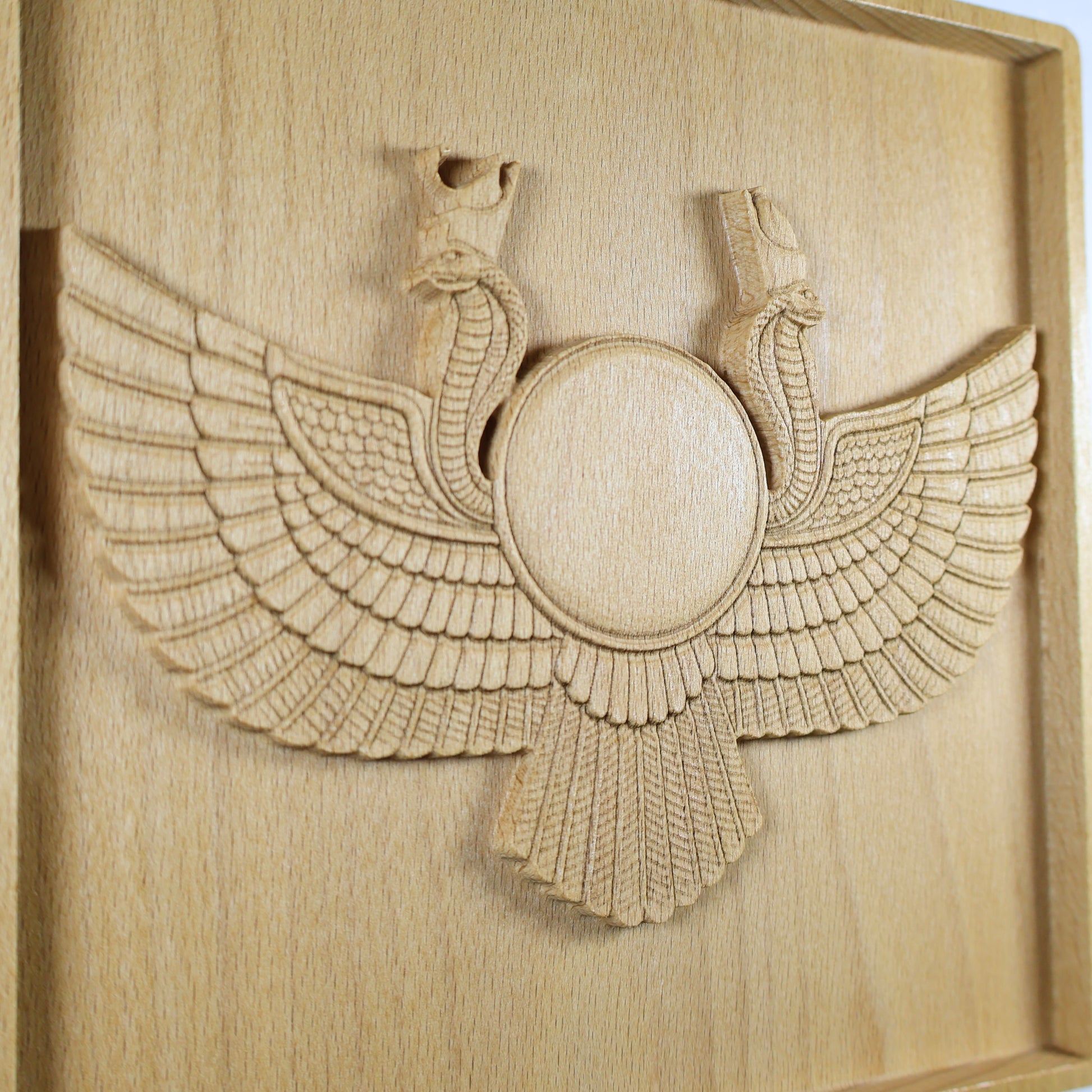 Angled close-up of an Egyptian Winged Sun Disk wood relief showing the Ra-Horus sun emblem, cobra forms, layered feathers, fan-shaped lower wing, and natural beech wood finish.