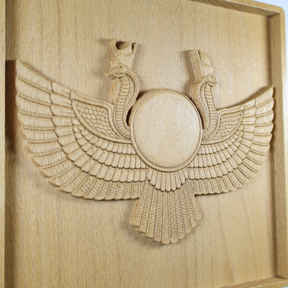 Angled close-up of an Egyptian Winged Sun Disk wood relief showing the Ra-Horus sun emblem, cobra forms, layered feathers, fan-shaped lower wing, and natural beech wood finish.
