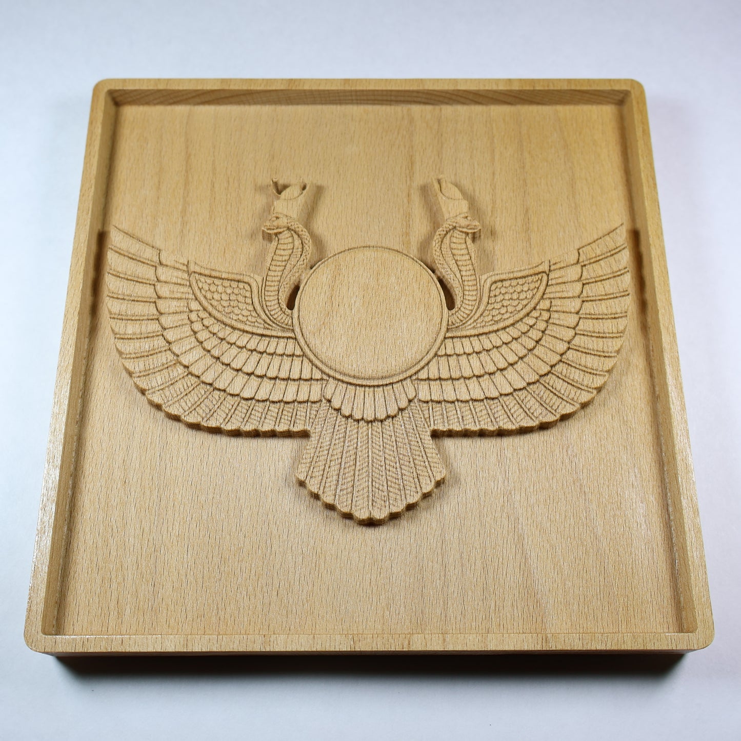 Angled view of an Egyptian Winged Sun Disk wood relief with a central sun disk, twin cobra details, layered wings, raised carving depth, and natural wood finish.