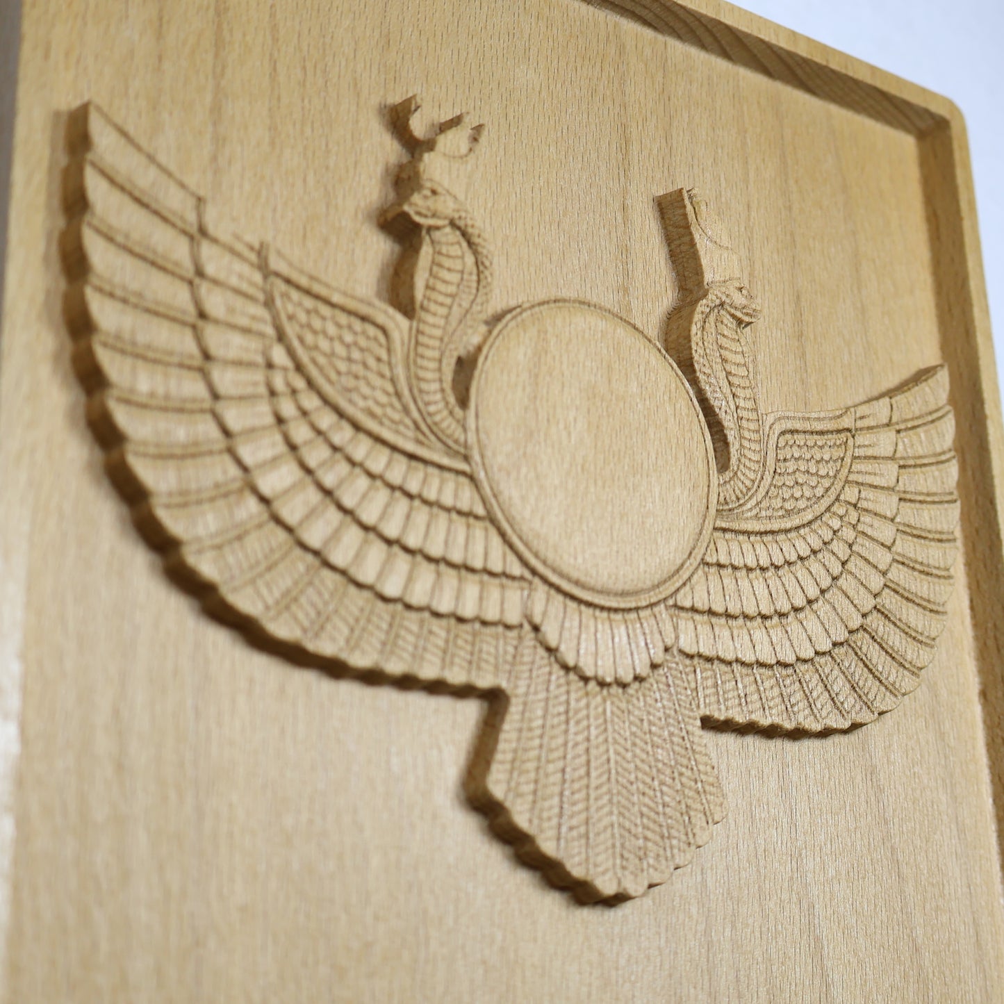Close angled detail of an Egyptian Winged Sun Disk wood relief showing the central sun disk, twin cobras, feathered wings, lower tail, carved depth, and natural beech wood finish.
