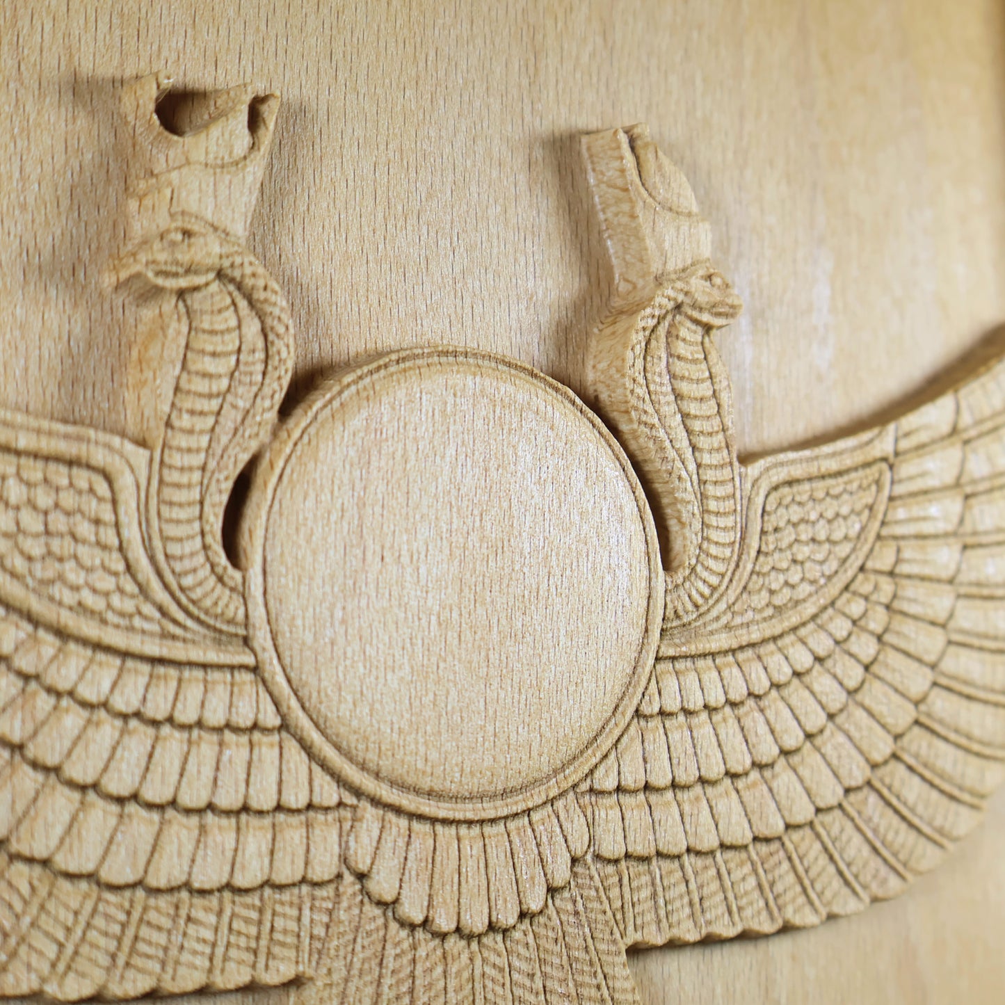 Macro detail of an Egyptian Winged Sun Disk wood relief showing the raised sun circle, cobra heads, layered feather carving, and natural beech wood grain.