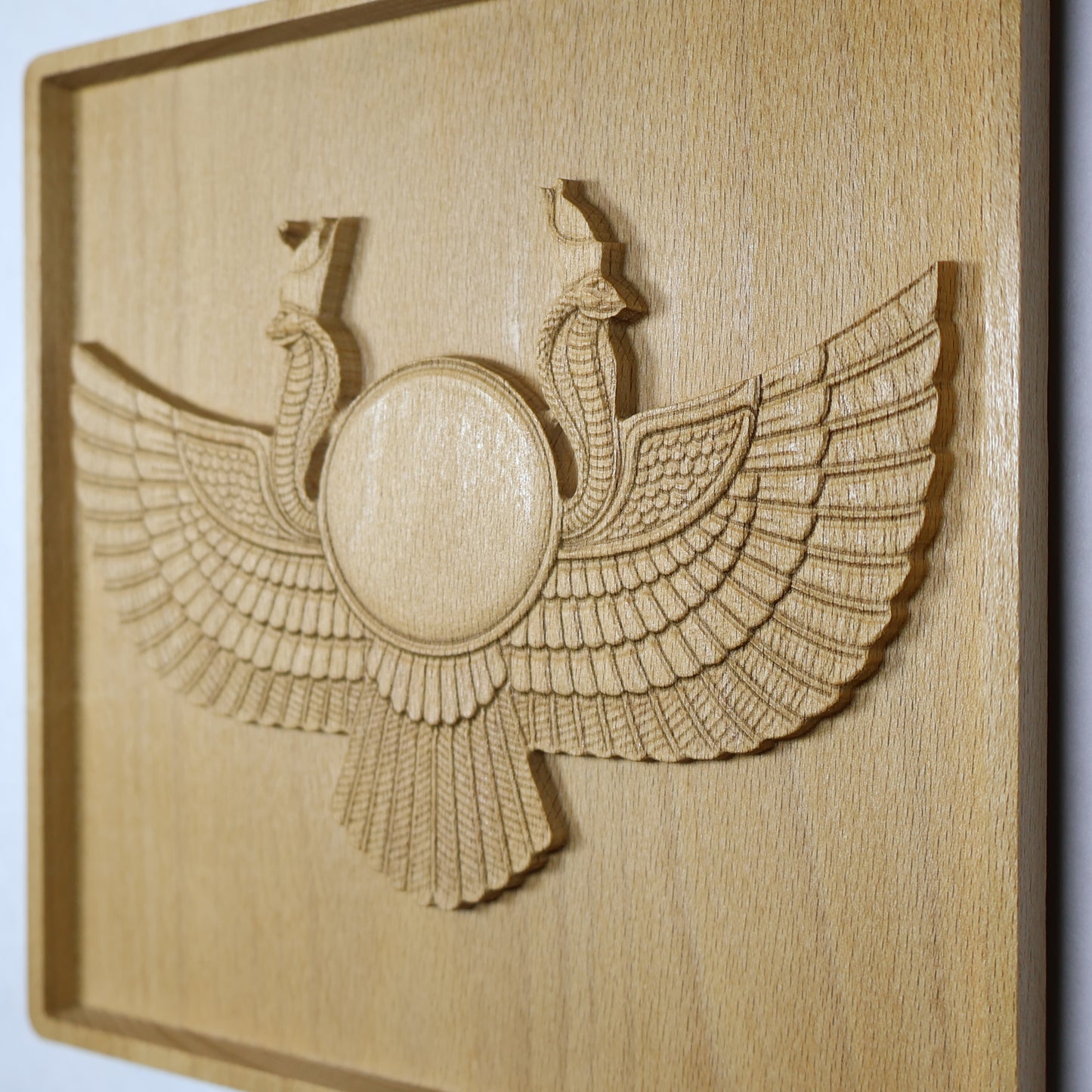 Display-angle view of an Egyptian Winged Sun Disk wood relief showing the raised sun disk, twin cobra guardians, broad carved wings, shadowed edges, and natural beech wood finish.