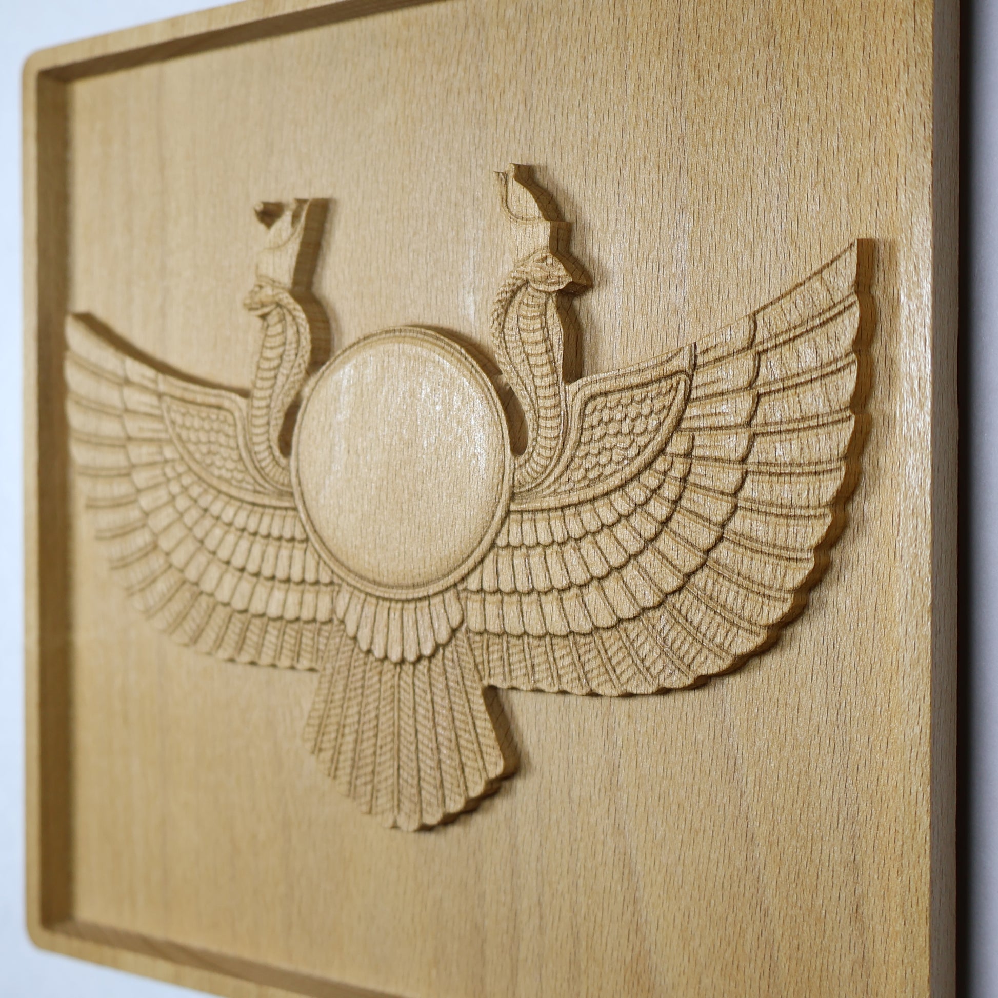 Display-angle view of an Egyptian Winged Sun Disk wood relief showing the raised sun disk, twin cobra guardians, broad carved wings, shadowed edges, and natural beech wood finish.