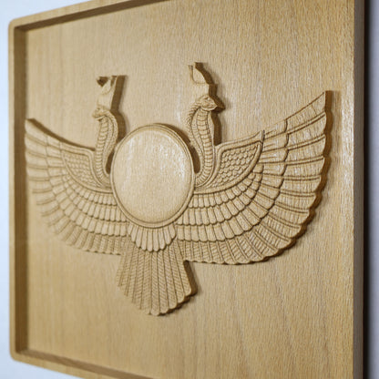 Display-angle view of an Egyptian Winged Sun Disk wood relief showing the raised sun disk, twin cobra guardians, broad carved wings, shadowed edges, and natural beech wood finish.