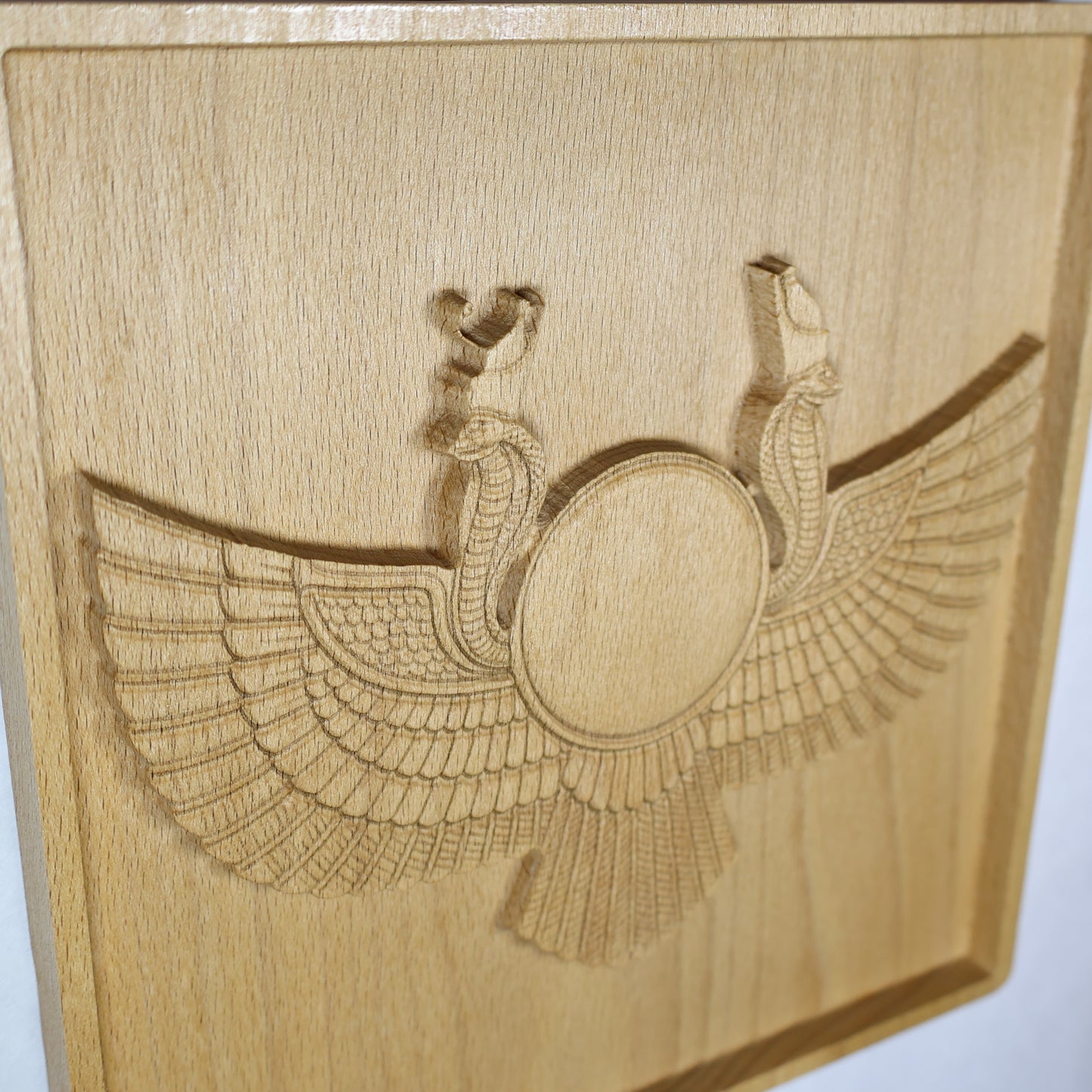 High side-angle view of an Egyptian Winged Sun Disk wood relief showing the raised sun emblem, cobra guardians, sweeping carved wings, recessed frame, and natural beech wood finish.