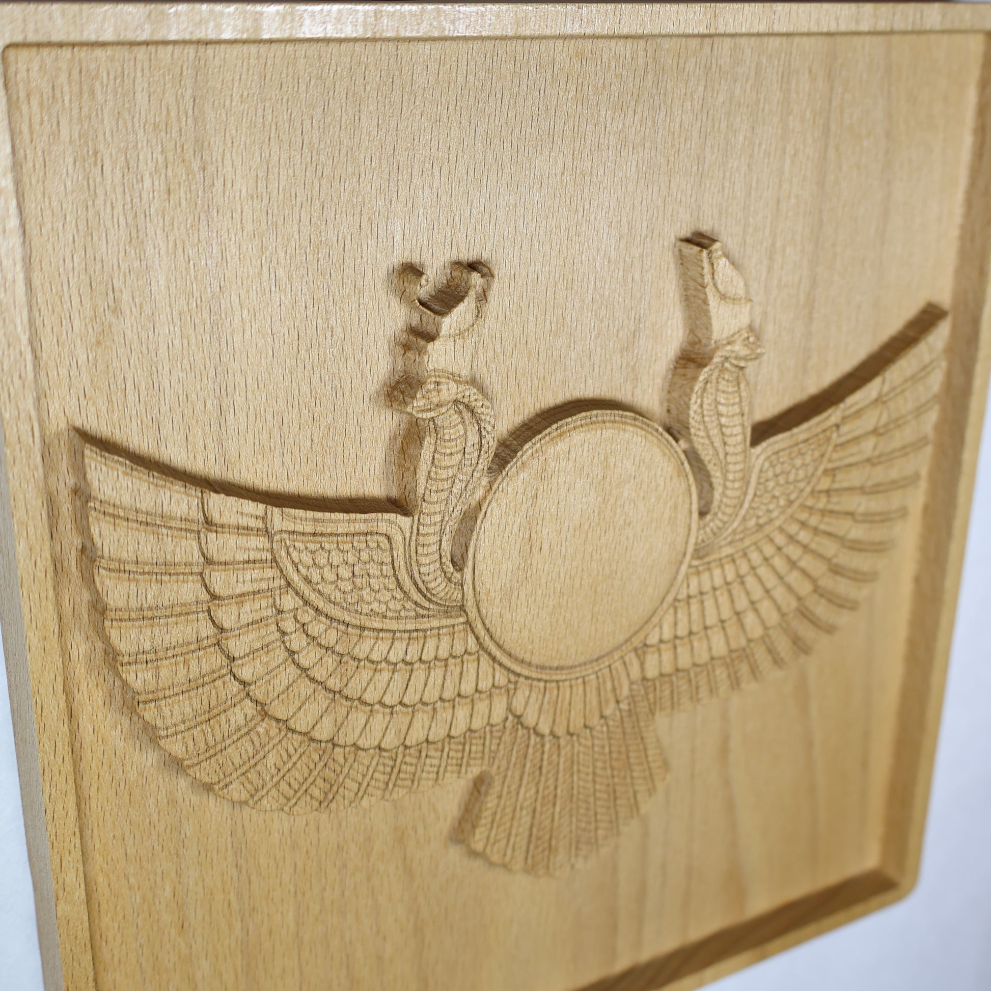 High side-angle view of an Egyptian Winged Sun Disk wood relief showing the raised sun emblem, cobra guardians, sweeping carved wings, recessed frame, and natural beech wood finish.