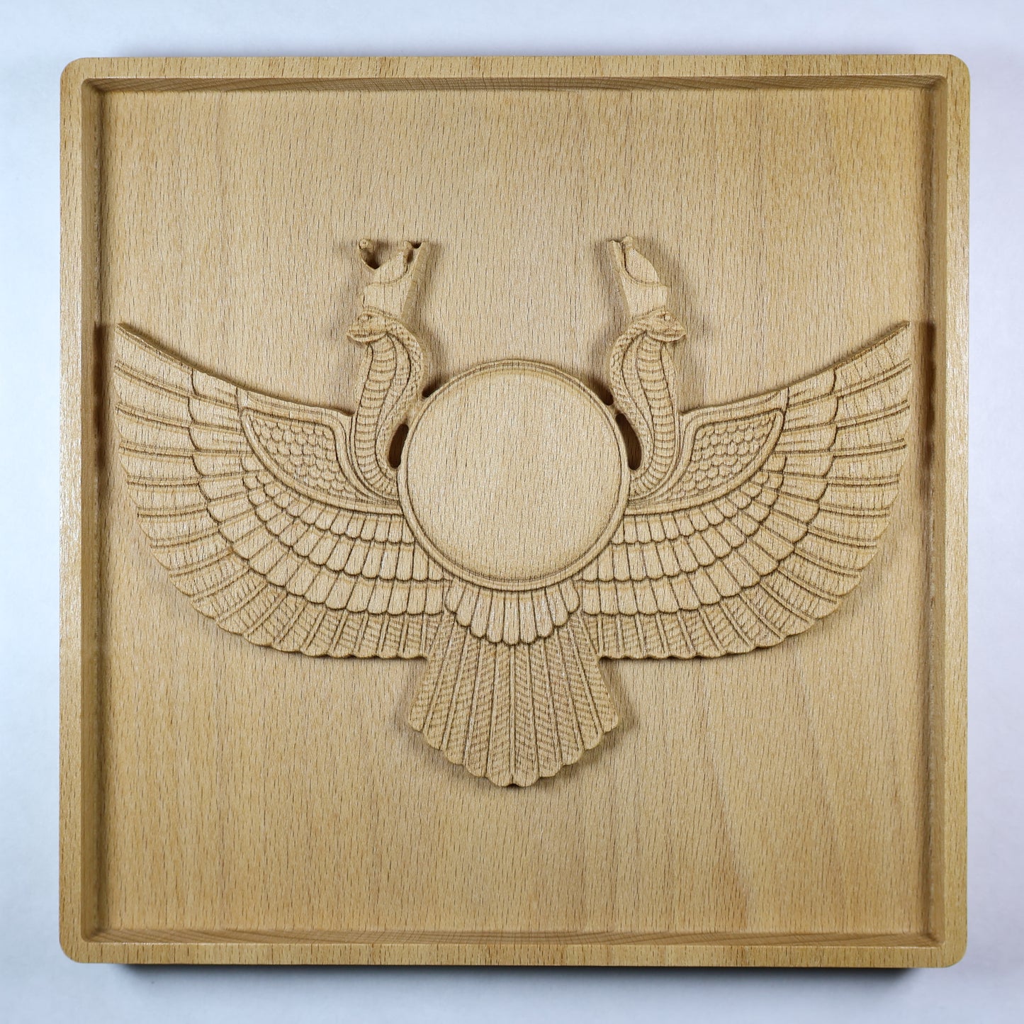 Straight-on view of an Egyptian Winged Sun Disk wood relief with a central sun disk, outstretched wings, paired cobra details, square frame, and natural wood finish.