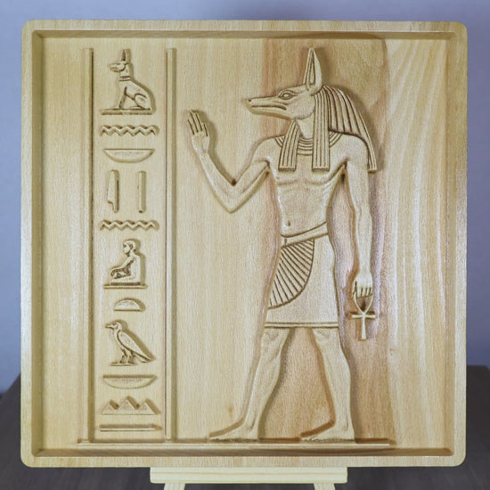 Rotating tabletop video of an Anubis wood relief, showing the front and angled side views of the Egyptian jackal-headed god, raised hand, ankh, hieroglyphic-style carvings, relief depth, and natural beech wood finish.